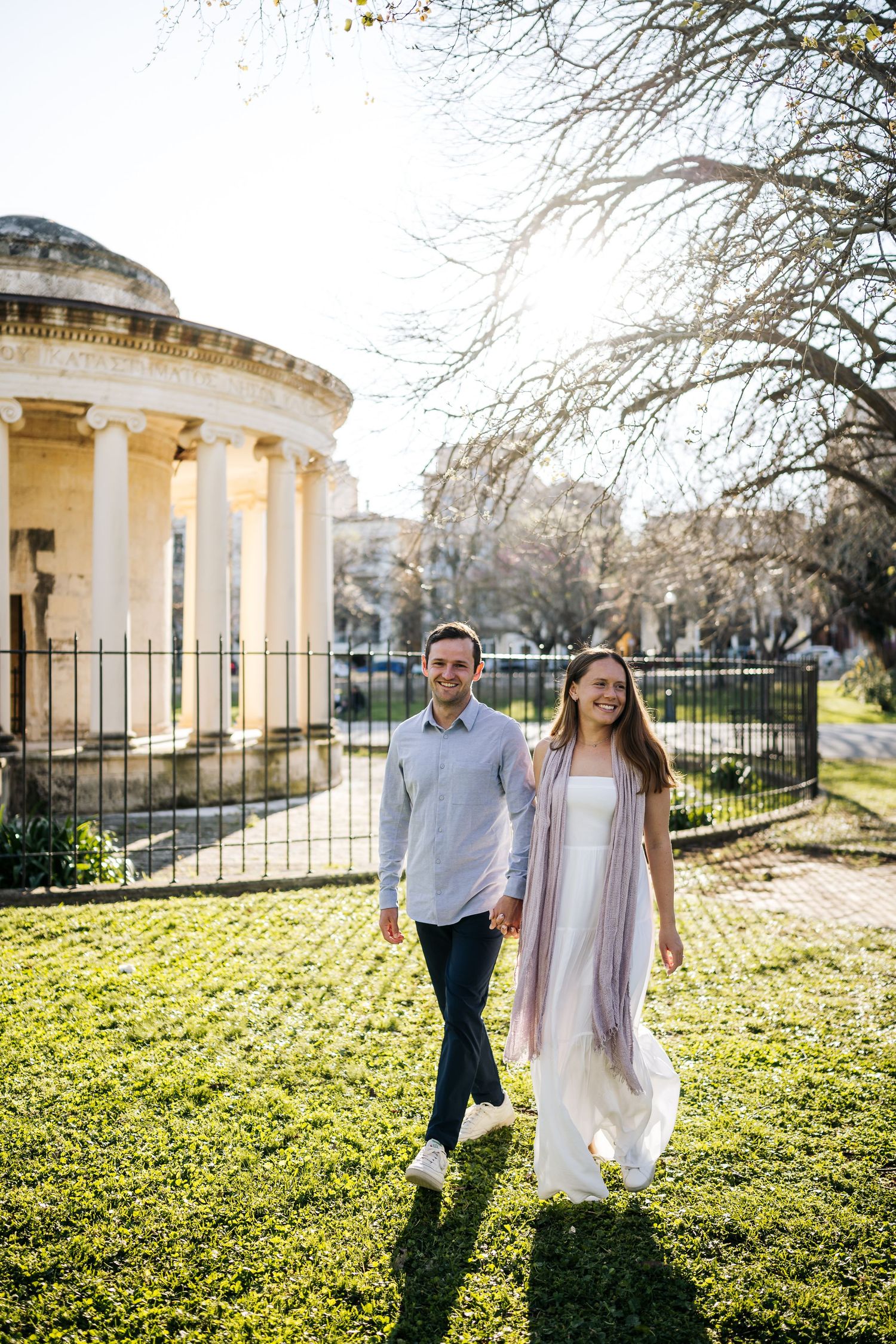A couple strolls through a sunny garden with neoclassical columns and architecture in the background.