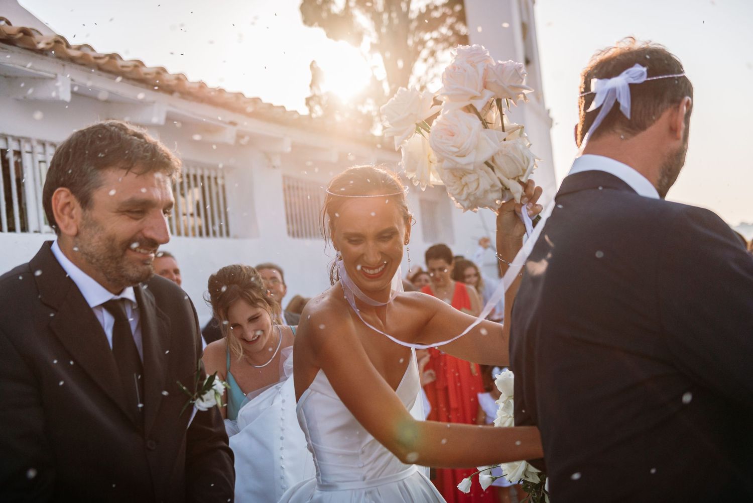 Sun-drenched wedding celebration with guests enjoying champagne against white building backdrop.