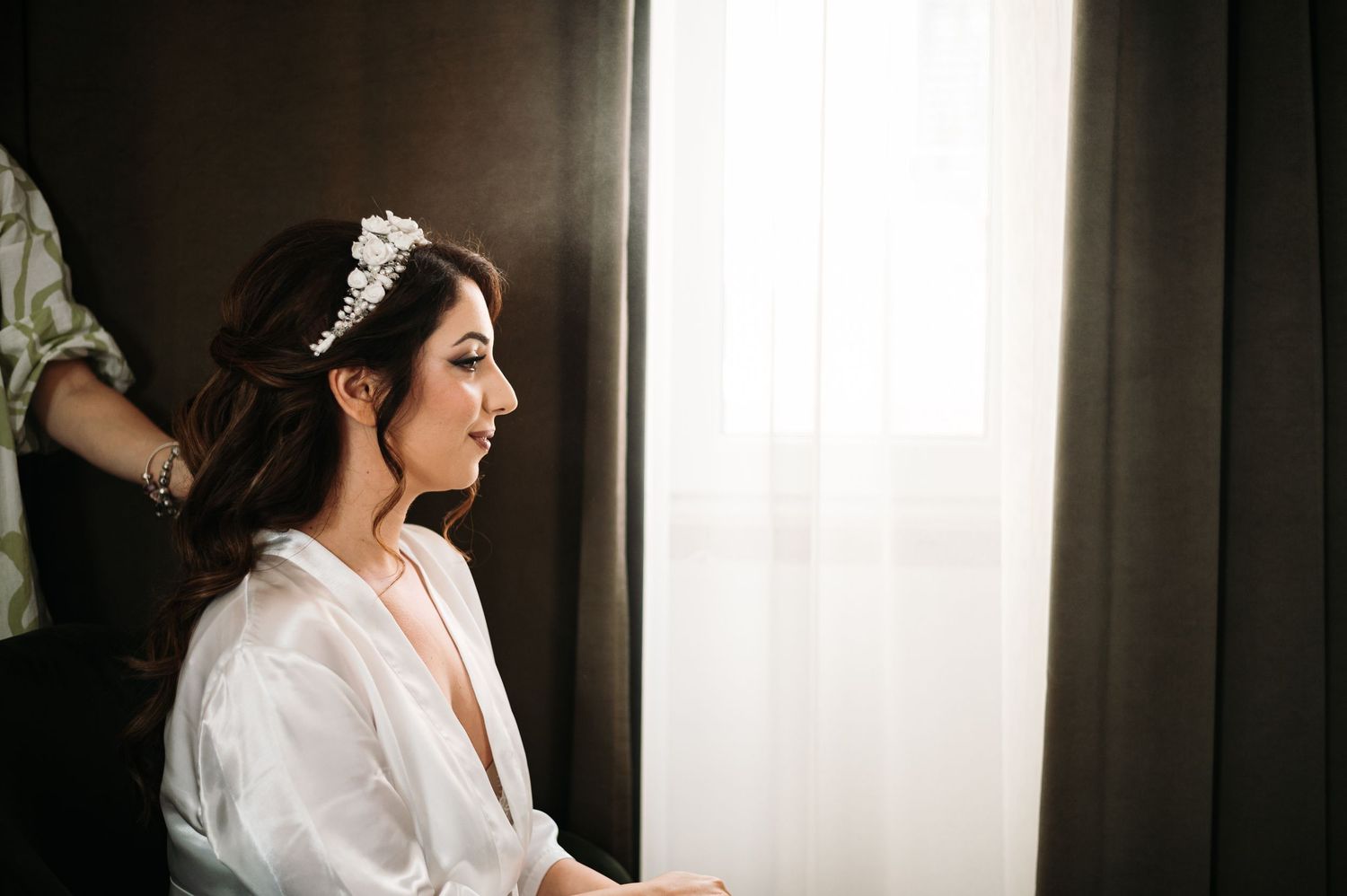 A person in a white robe sits by a window during wedding preparations.