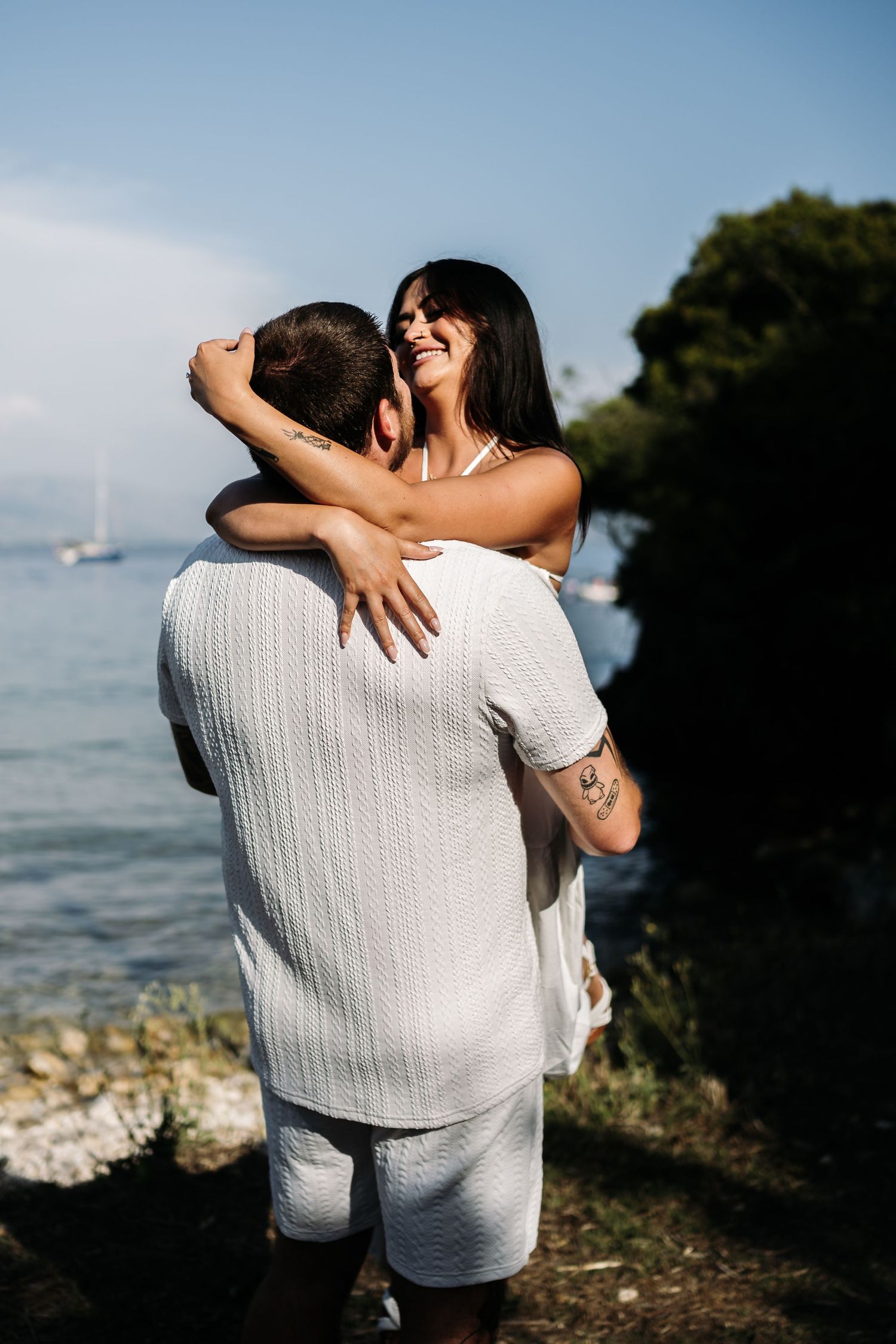 A romantic embrace by the ocean with a white-clad couple sharing a joyful moment against a coastal backdrop.
