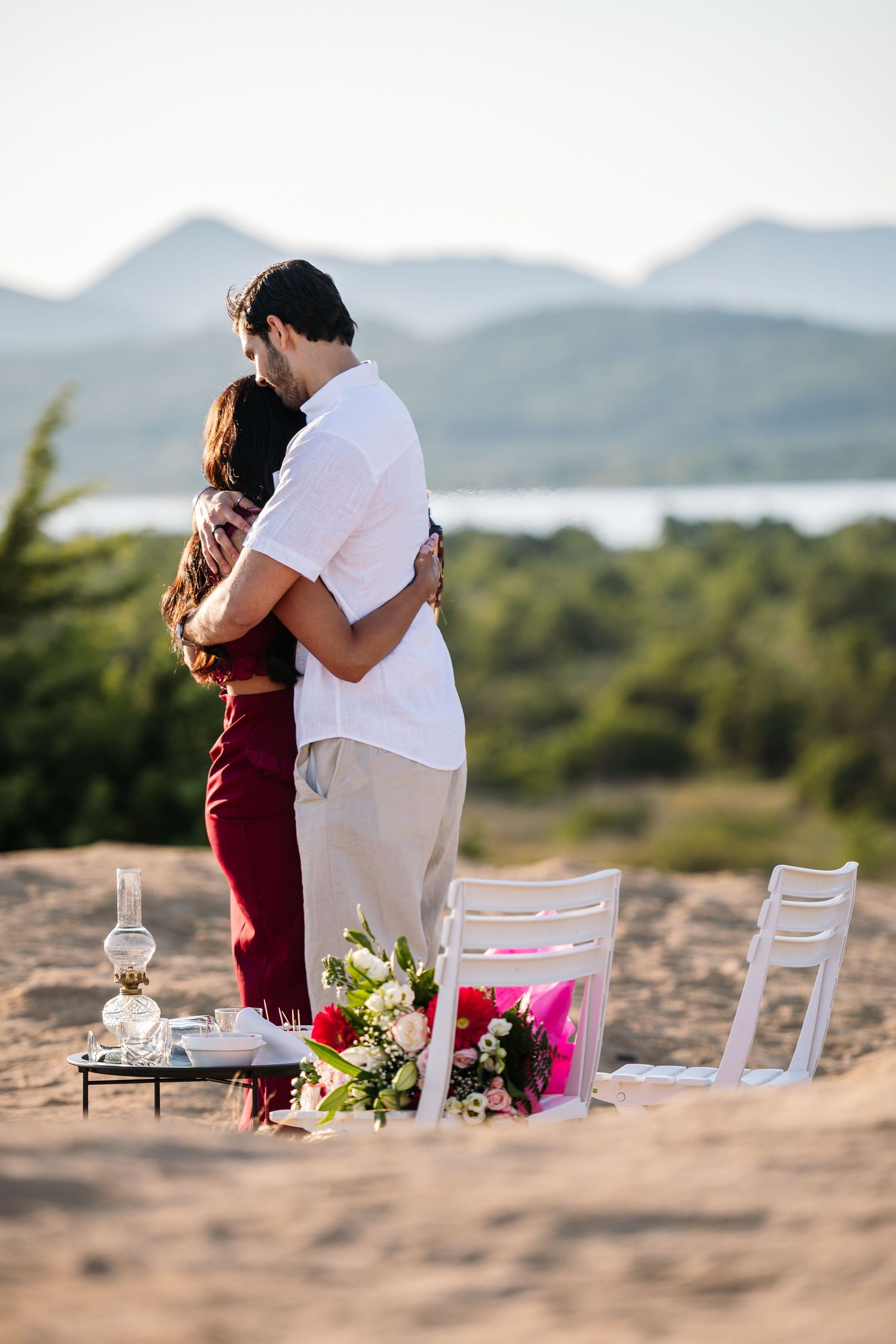Romantic beach setup with white chairs and floral arrangements against mountain backdrop.