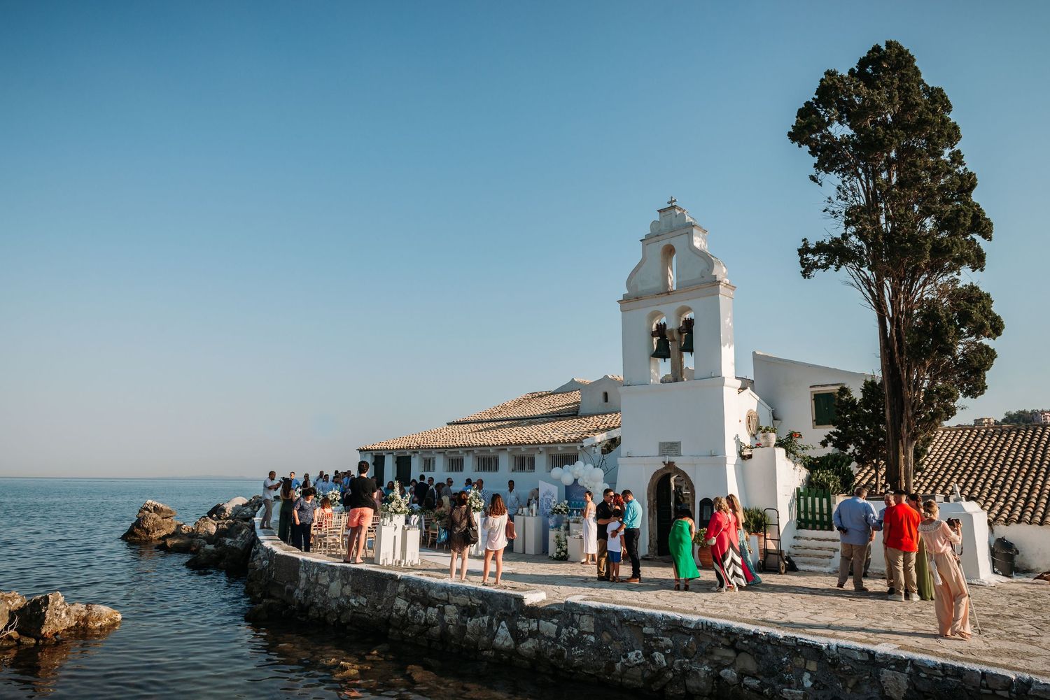 Mediterranean seaside wedding venue with white chapel and guests gathered by the water.