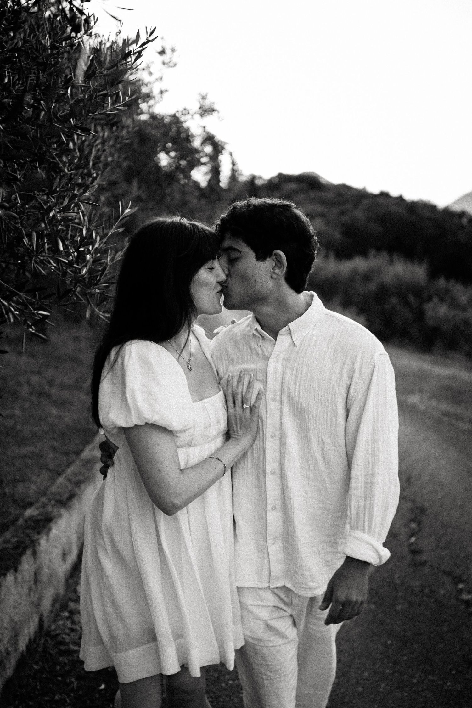 An intimate black and white photo of a couple in white clothing embracing on a countryside path.
