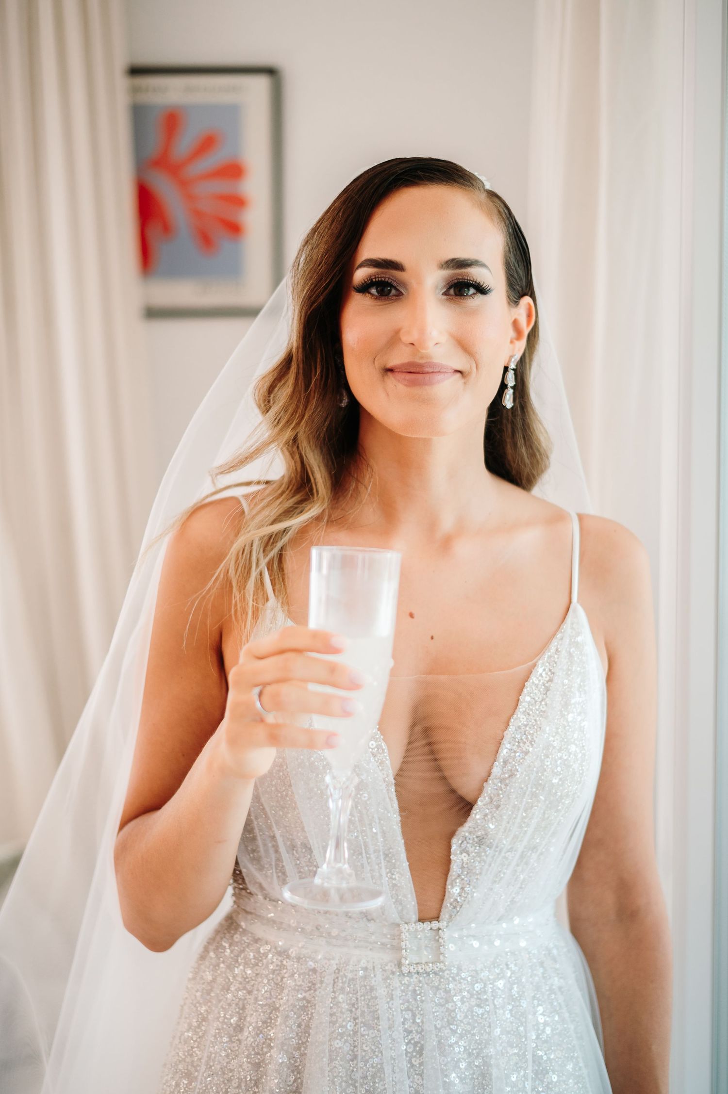 Bride in sparkling white dress holding champagne glass during wedding preparations.
