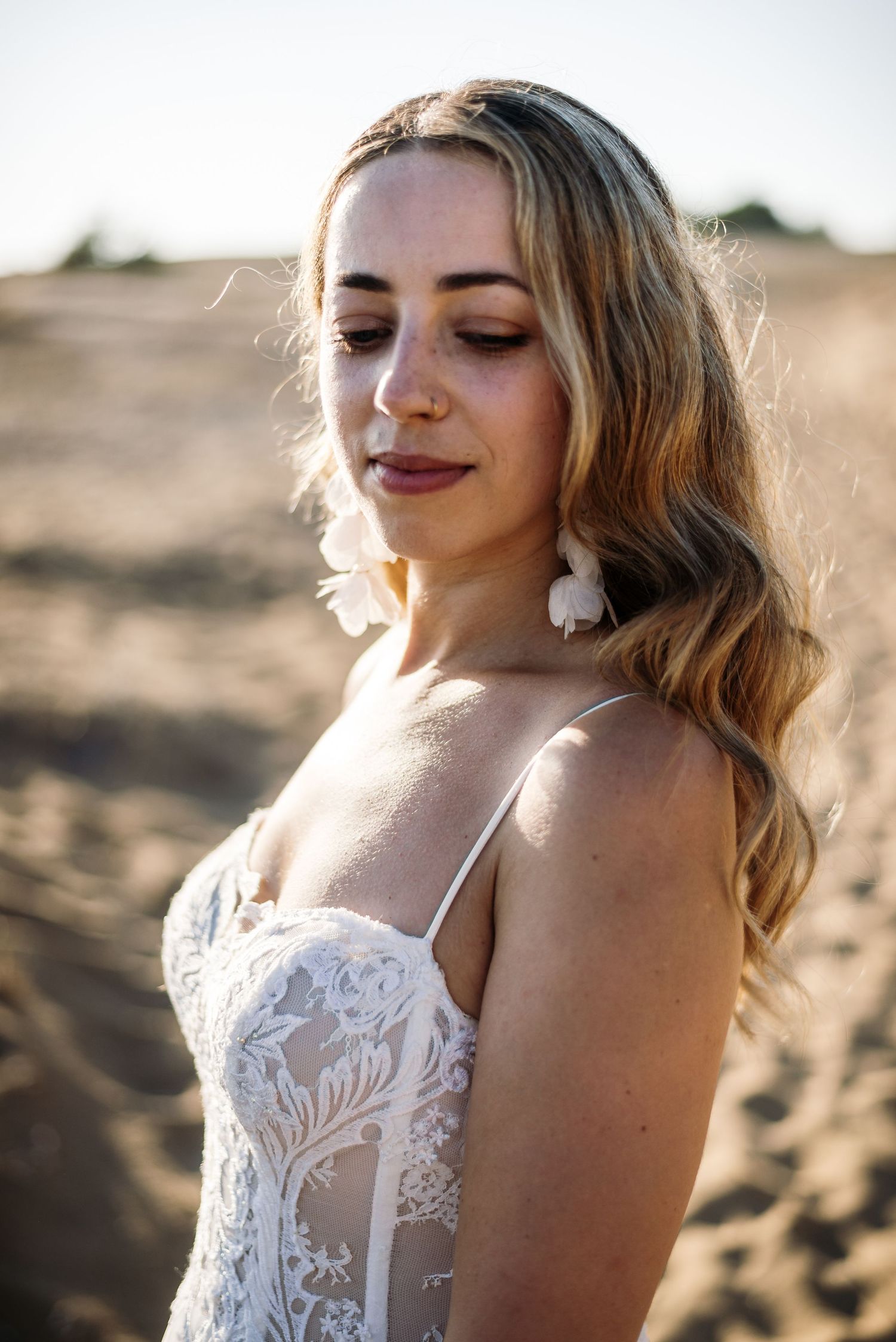 A bride in a detailed lace wedding dress poses in golden sunlight wearing statement earrings.