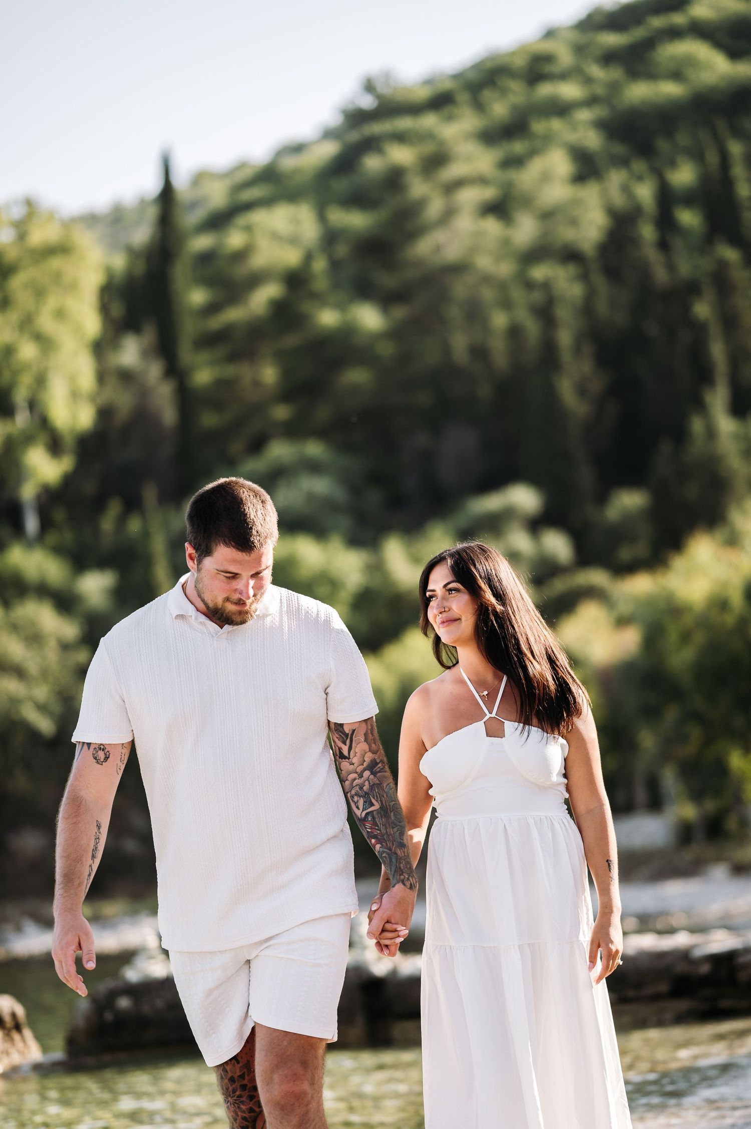 A couple in matching white outfits walking hand in hand along a Mediterranean coastline.