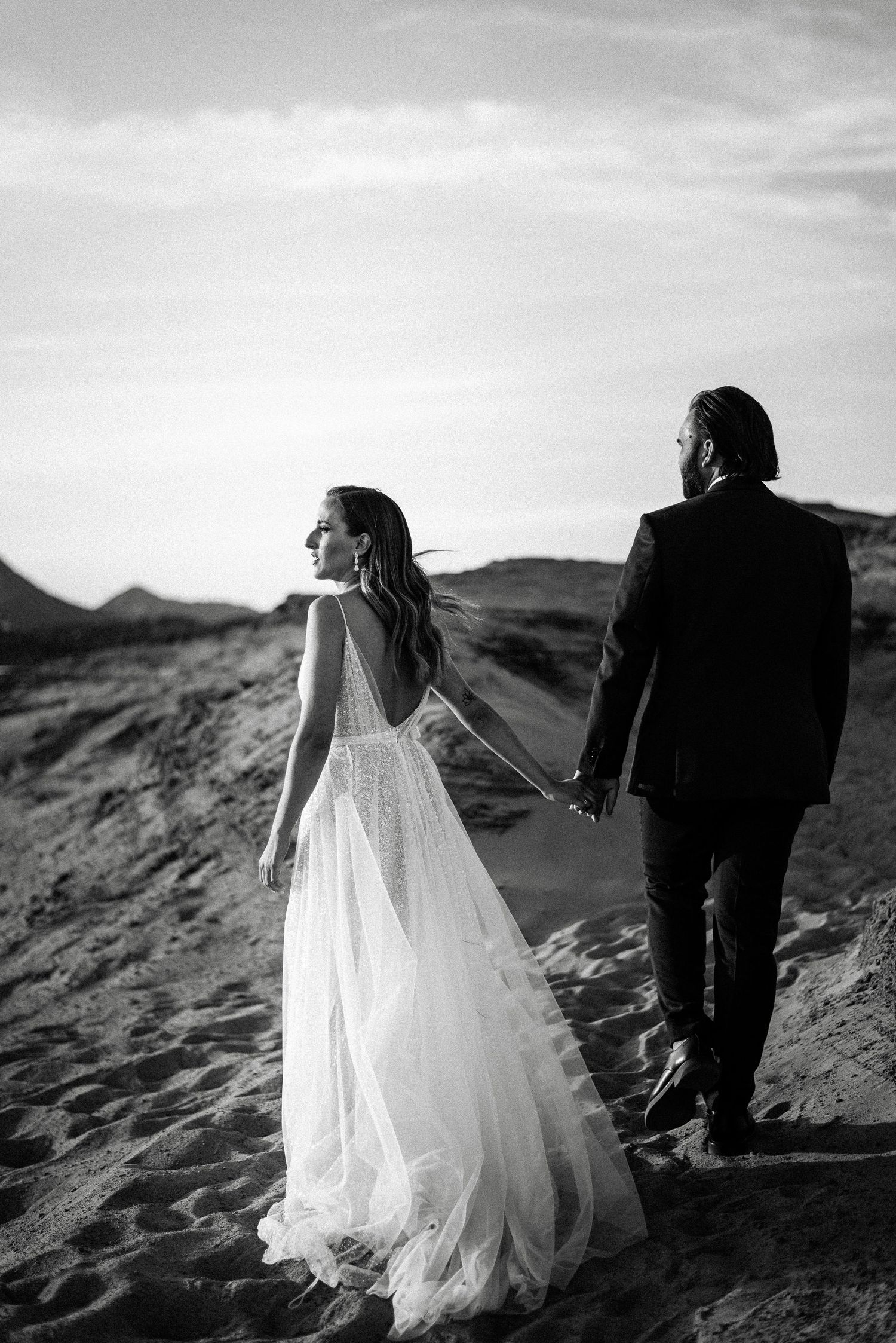 A couple in wedding attire walks along dramatic black and white coastal terrain.