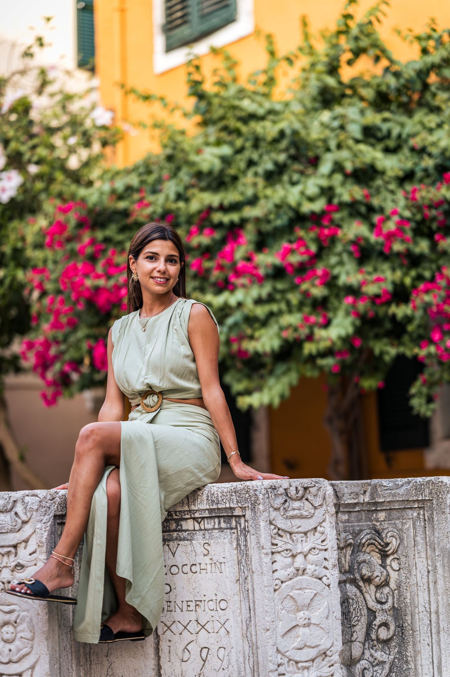 A woman in a mint green dress poses on stone steps near yellow walls and pink flowers.