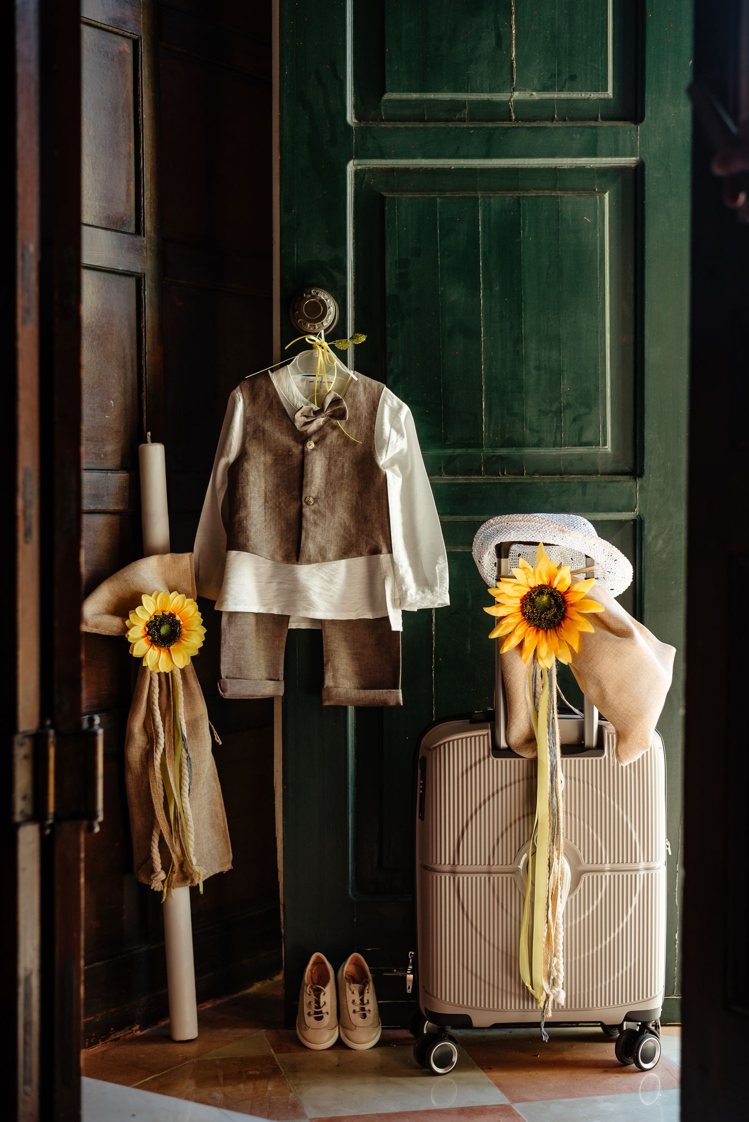 Formal children's outfit display with sunflowers against dark green door and luggage.