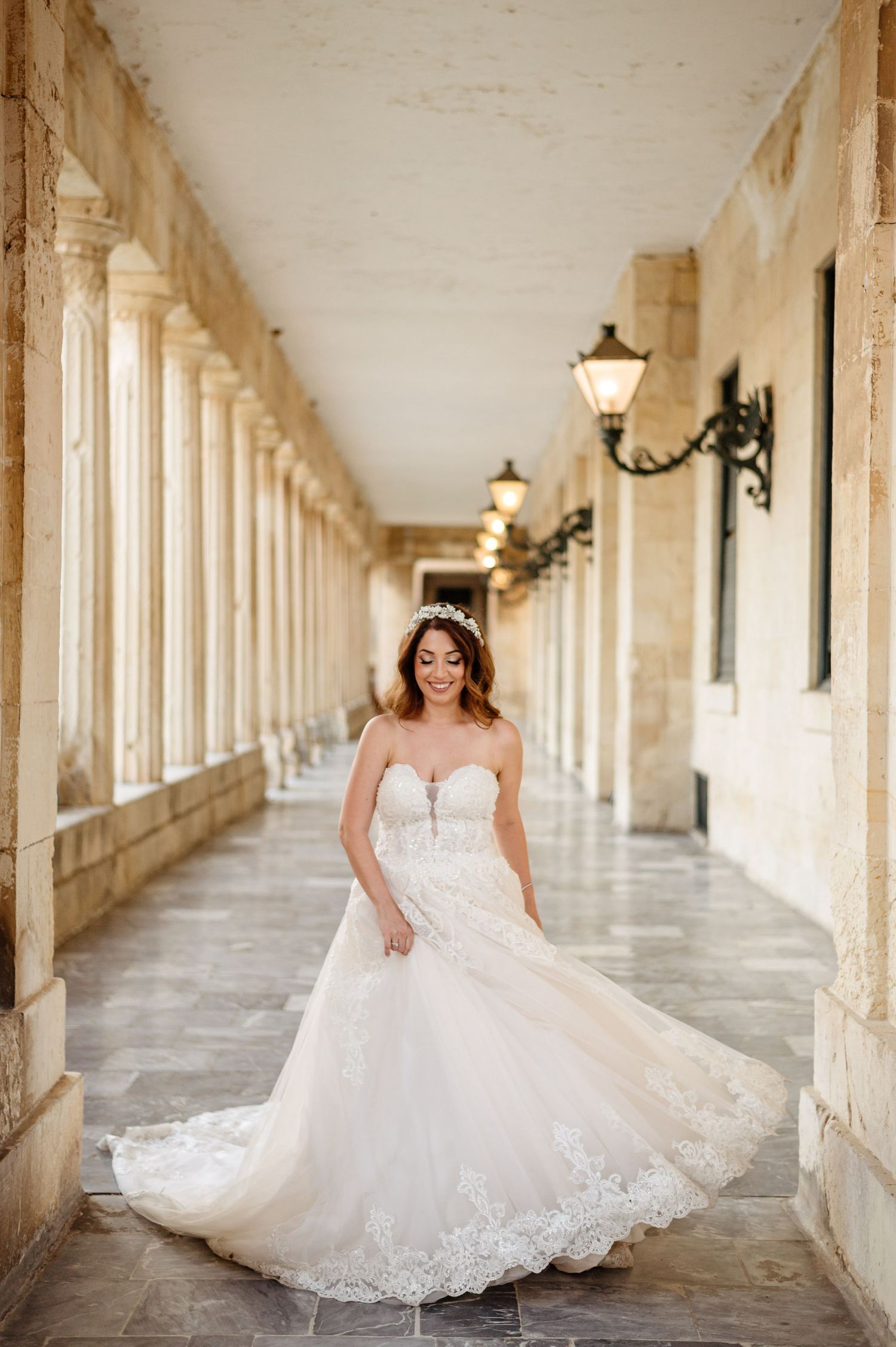 A bride in a lace wedding gown poses in an elegant colonnade with ornate lanterns and classical architecture.
