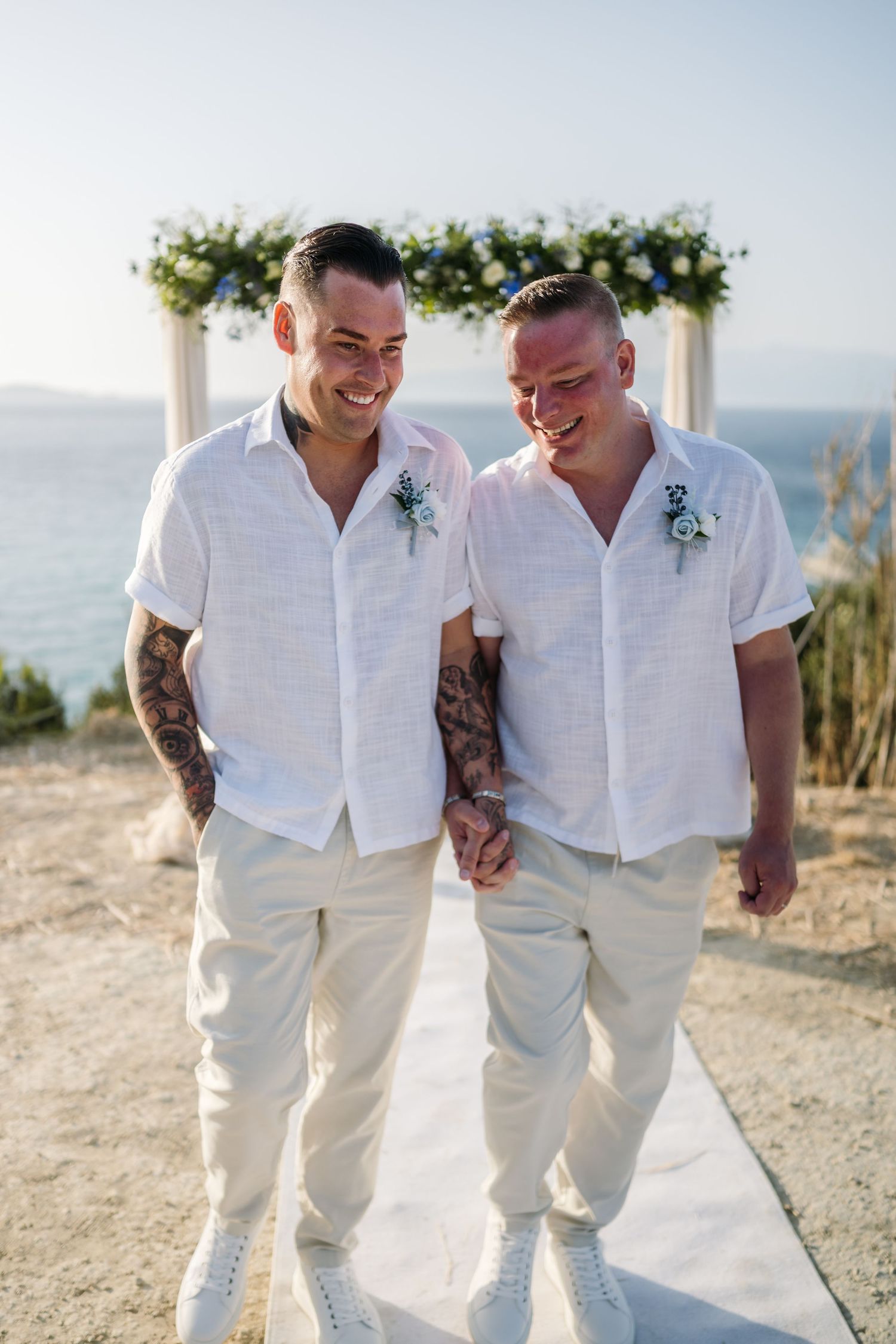 Happy couple in matching white attire sharing joyful moment under floral arch with ocean backdrop.