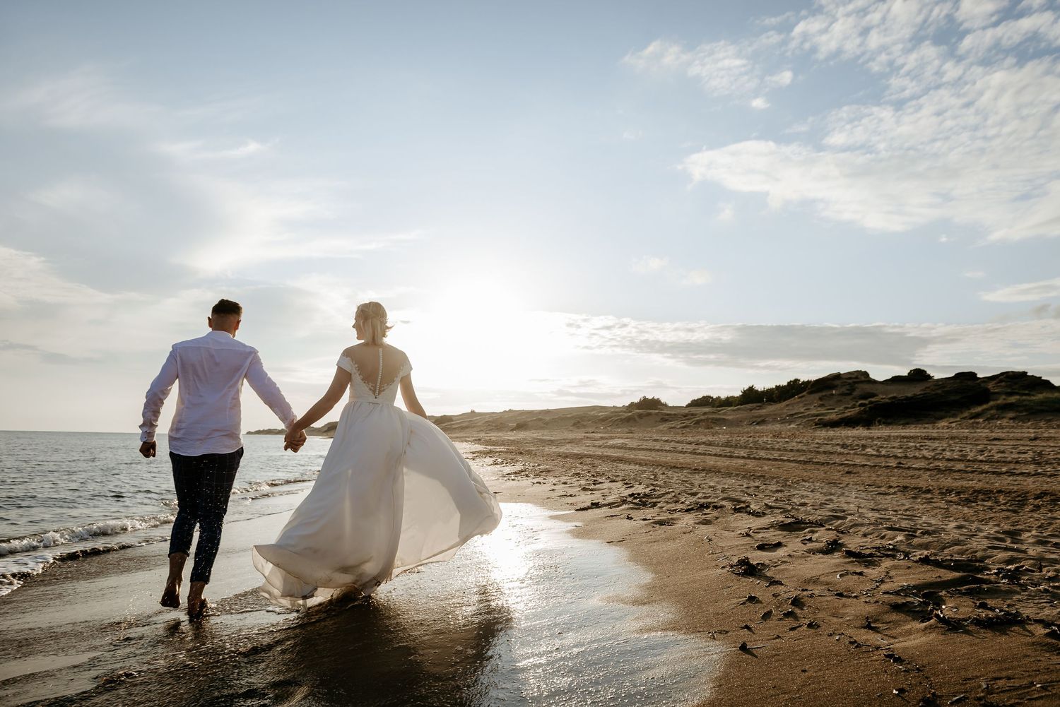 Couple walking along beach shoreline at sunset with flowing white dress.