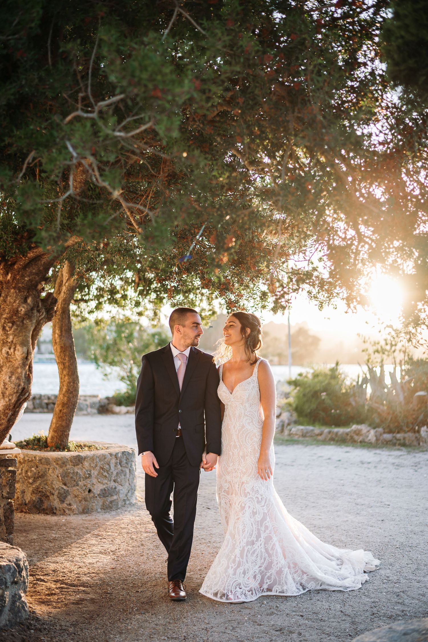 A romantic wedding portrait of a couple walking beneath a tree at sunset.