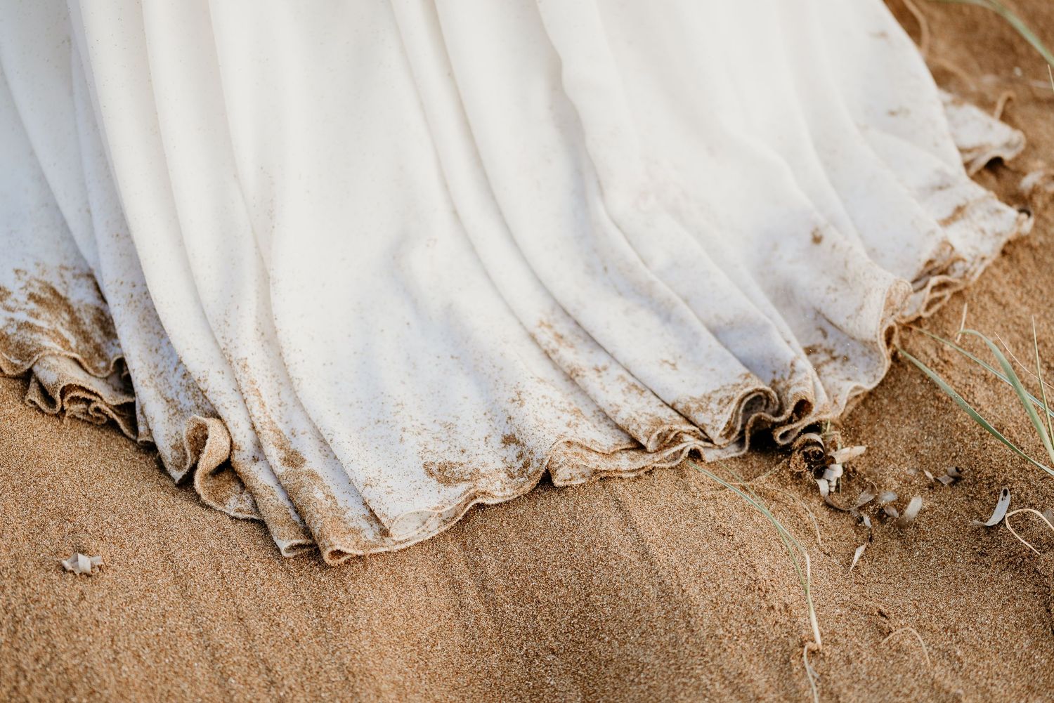 White wedding dress hem trailing in sand on a sunny beach.