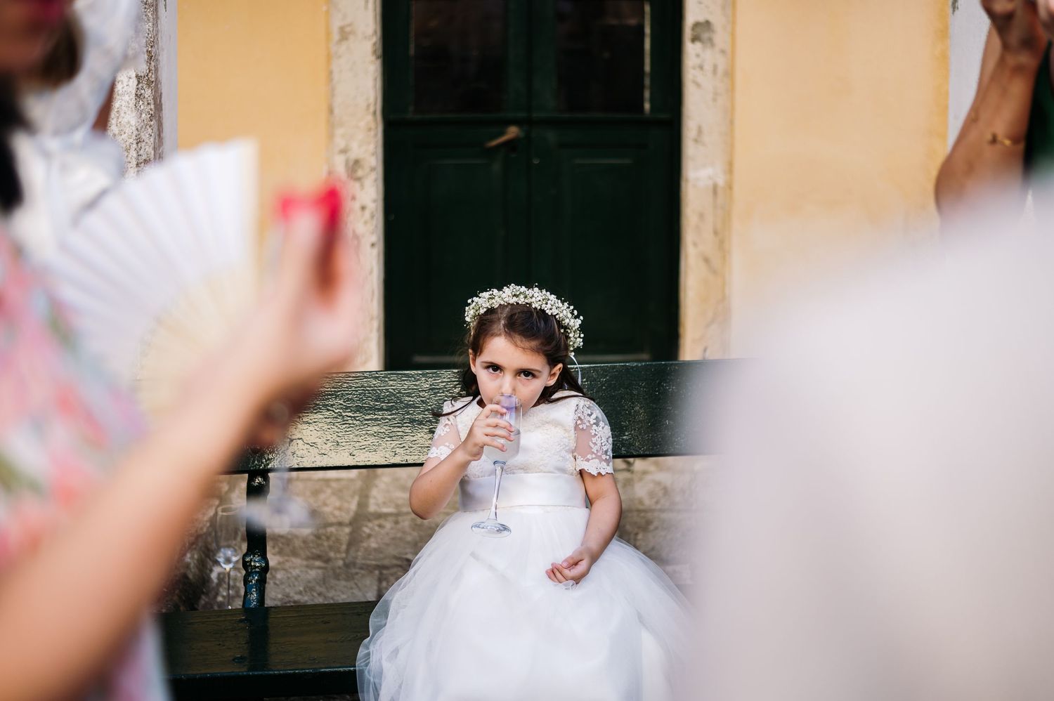 Young flower girl in white dress sips from a glass during wedding festivities.