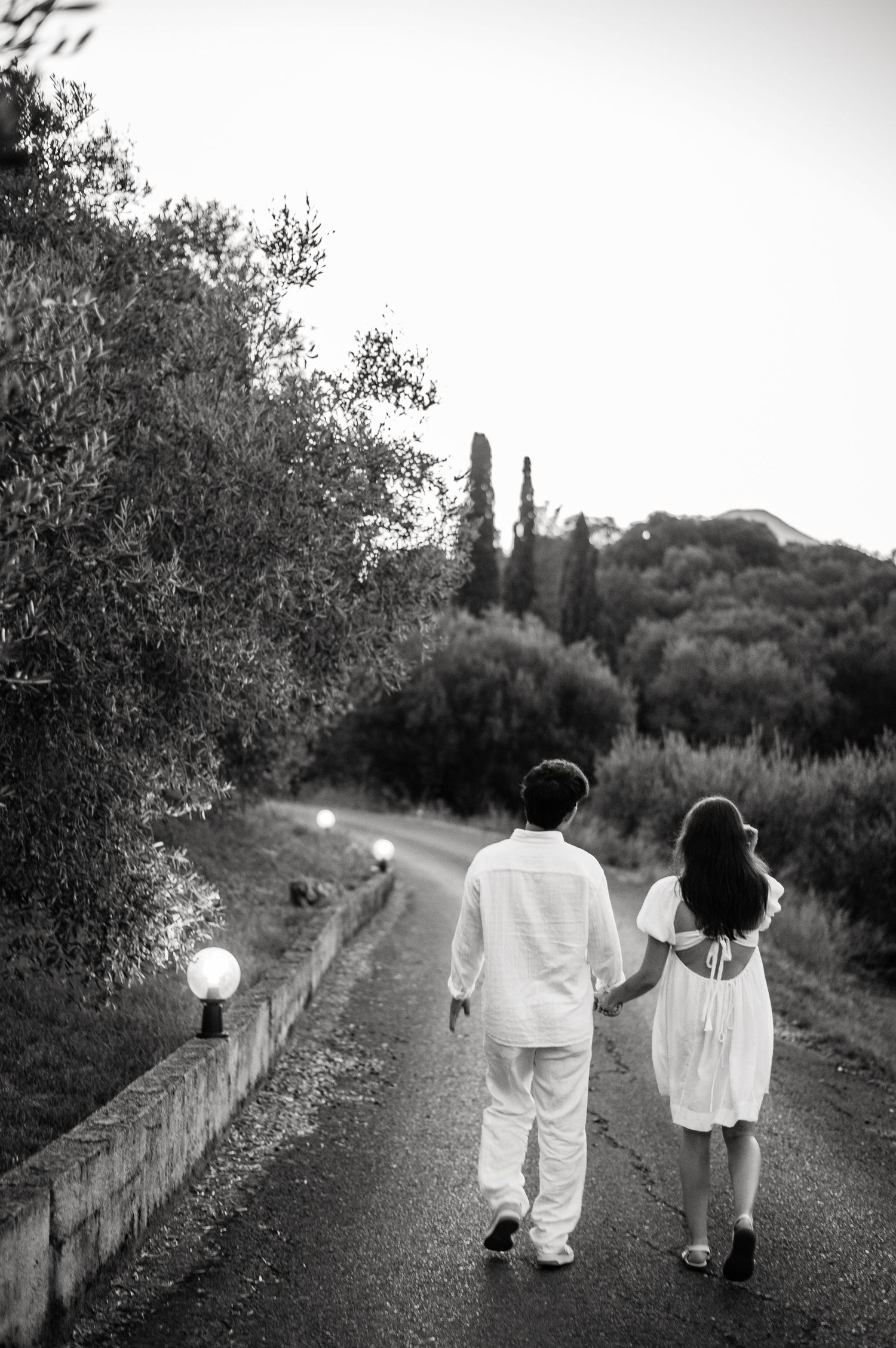 A couple in white outfits walking together on a scenic path surrounded by lush greenery and cypress trees.