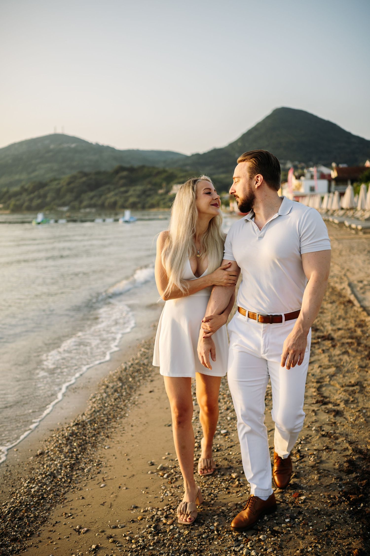 Couple in matching white outfits walking along a scenic beach with mountains in background.