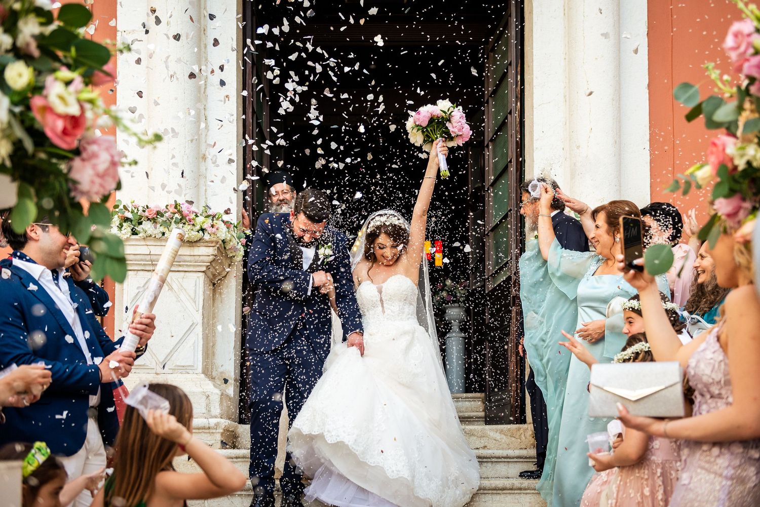Wedding guests throw confetti at the newlyweds outside a grand building entrance.