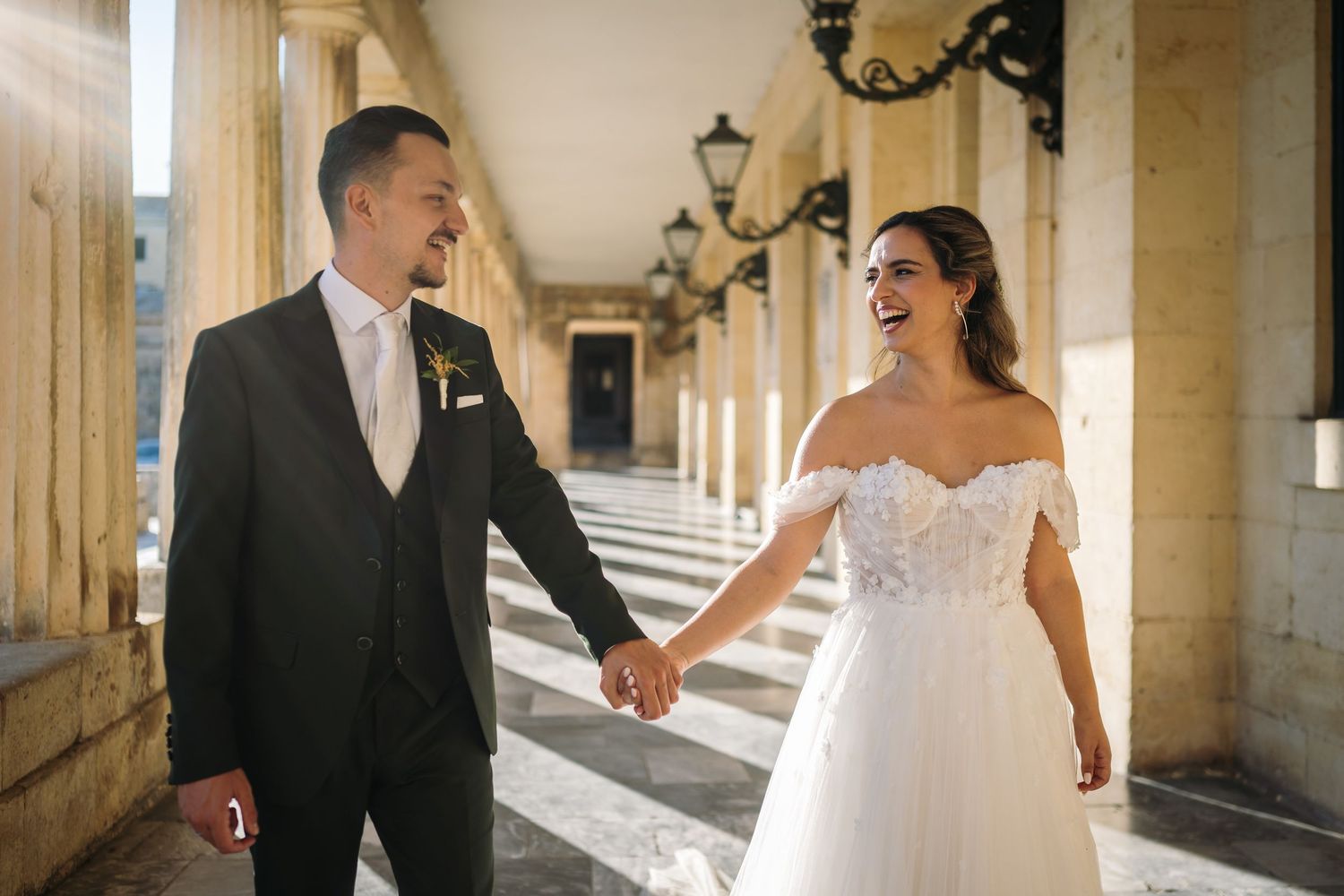 Wedding couple shares joyful moments in sunlit historic colonnade with classical architecture.