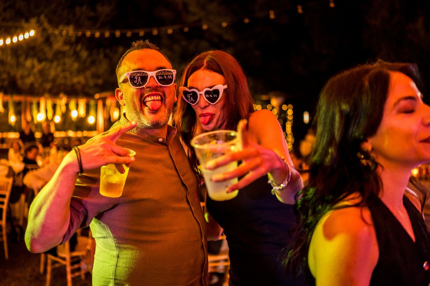 Party guests wearing neon sunglasses dance and celebrate at wedding reception.