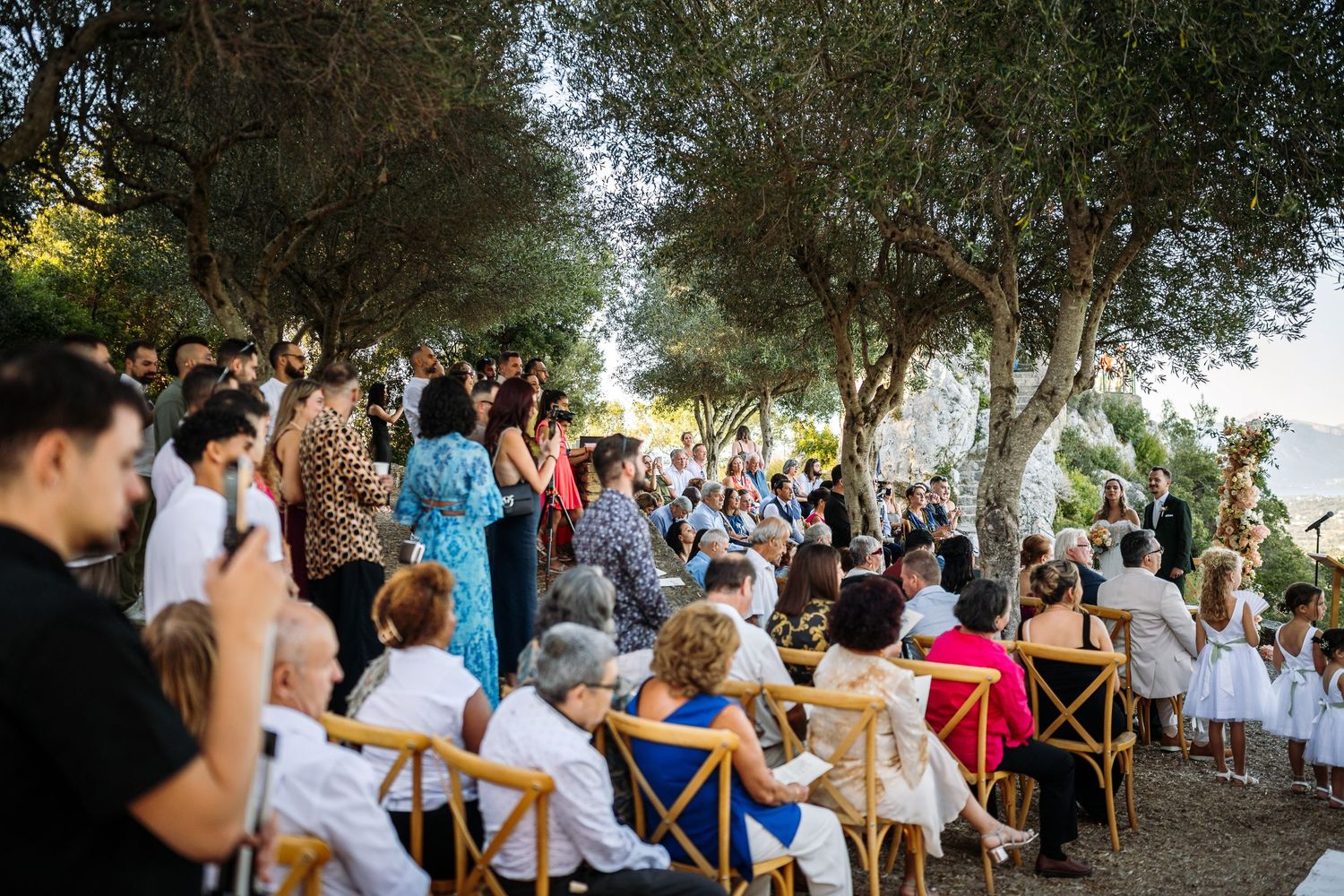 Wedding guests gather outdoors under trees for a celebration ceremony.