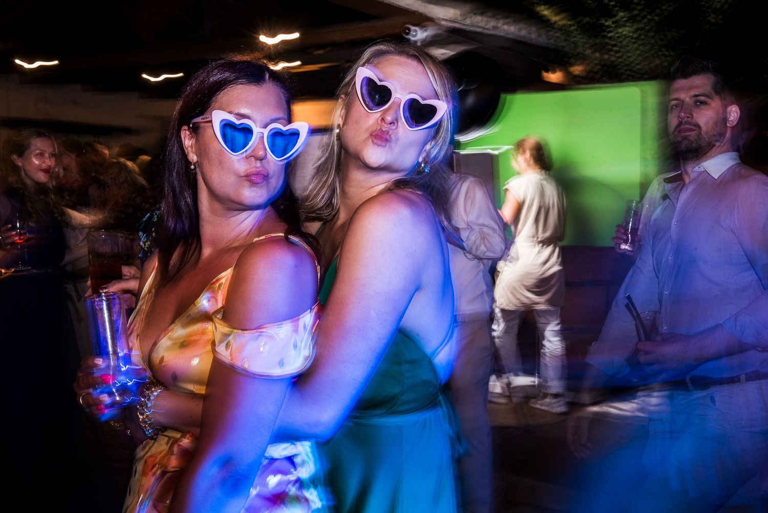 Party-goers wearing sunglasses dance under blue and purple lighting at a nighttime celebration.