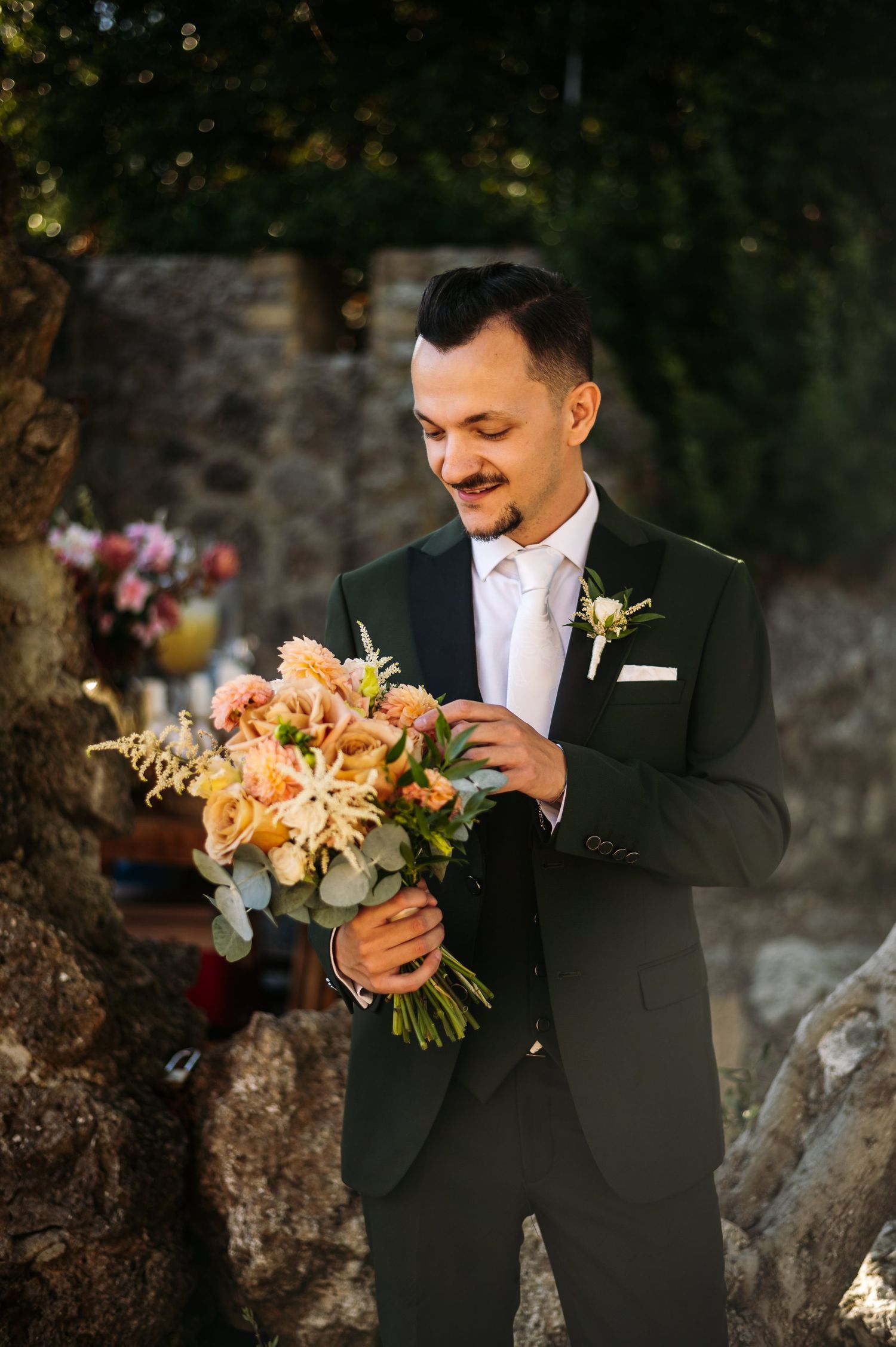 Person in formal black suit holds a peach and cream floral wedding bouquet outdoors.
