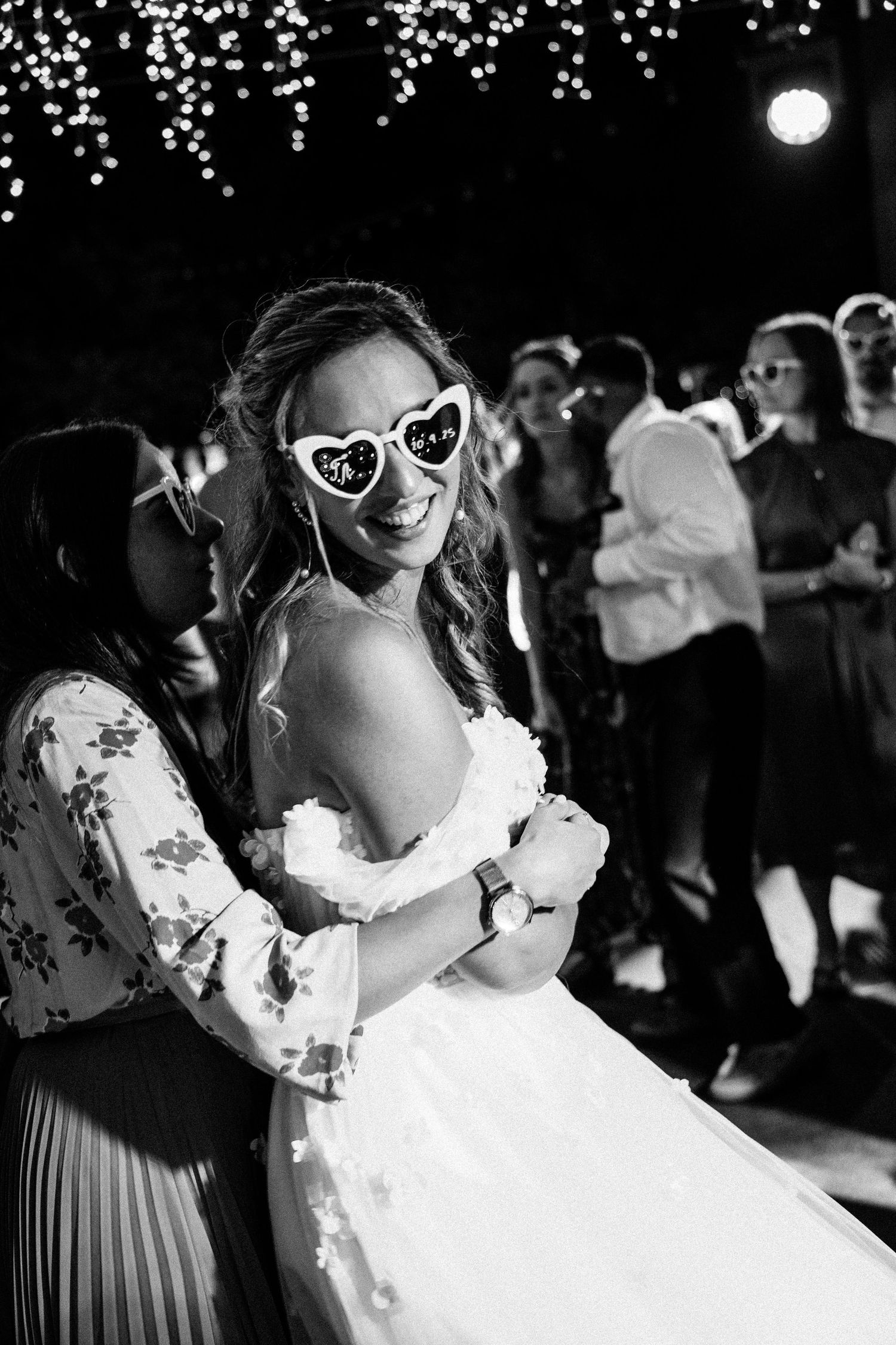 Black and white photo of wedding guests dancing under sparkling lights wearing sunglasses.