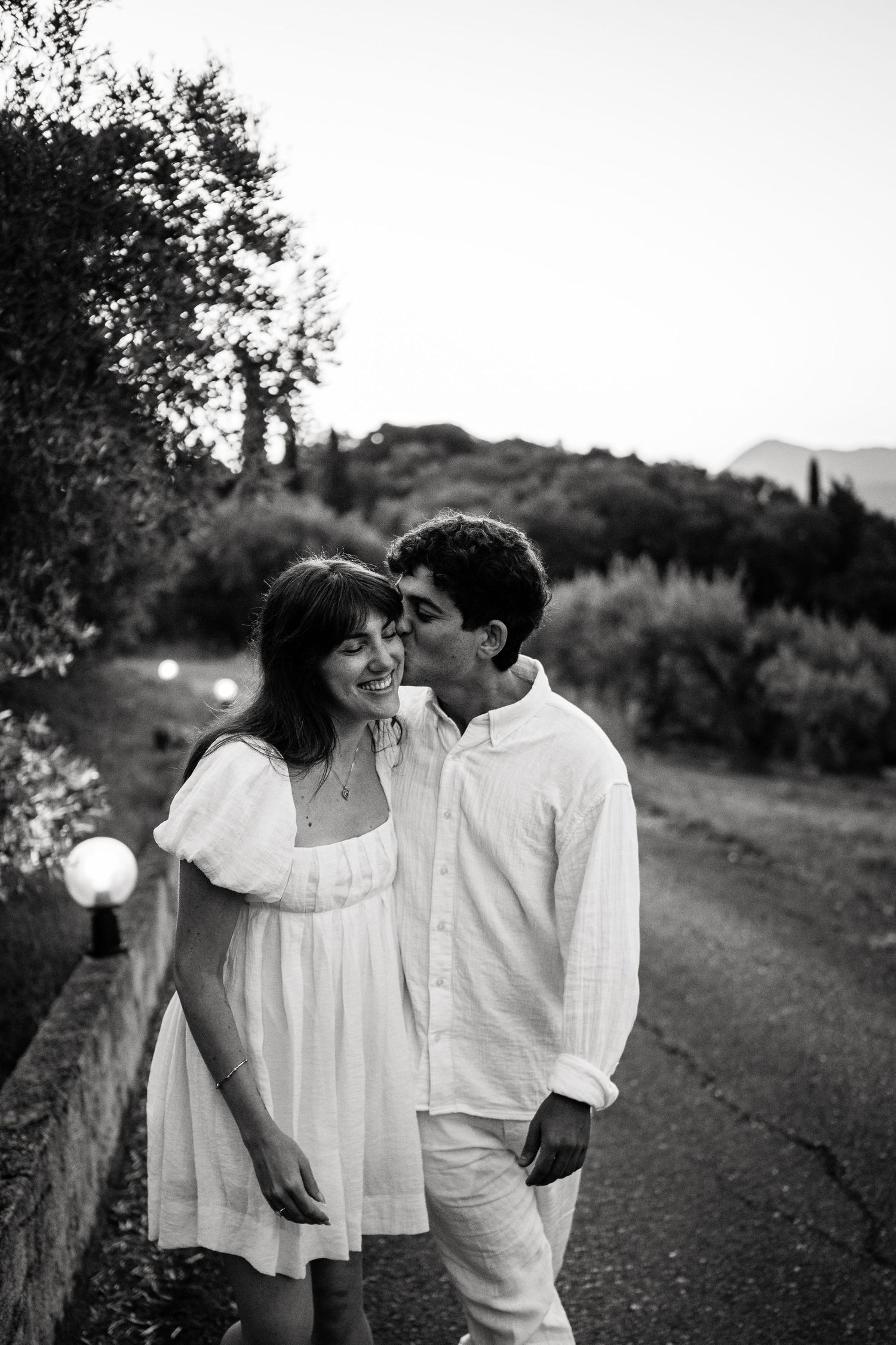 A romantic black and white portrait of a couple in white clothing sharing an intimate moment on a garden path.