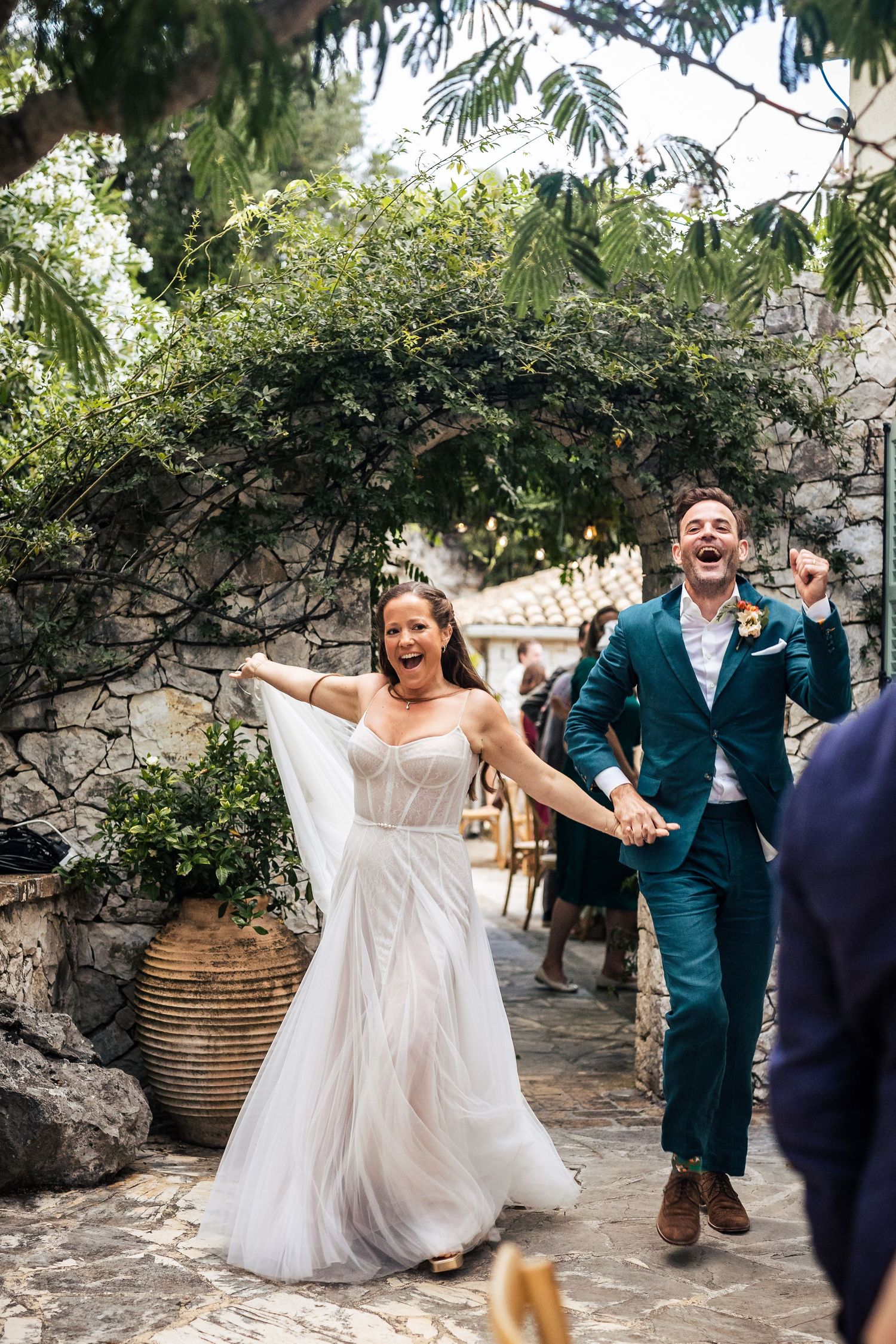 Wedding party dances and celebrates in a lush garden setting with stone walls.