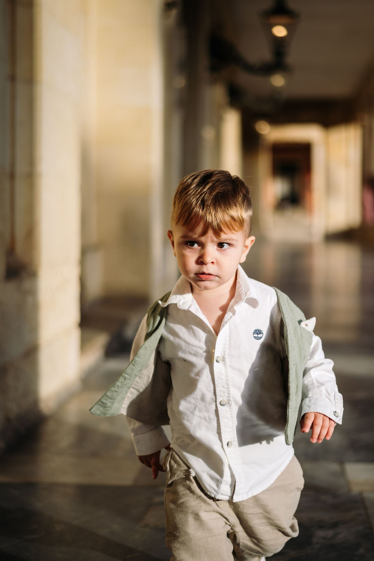 Young child in white shirt and khaki pants walking through an elegant hallway with columns.