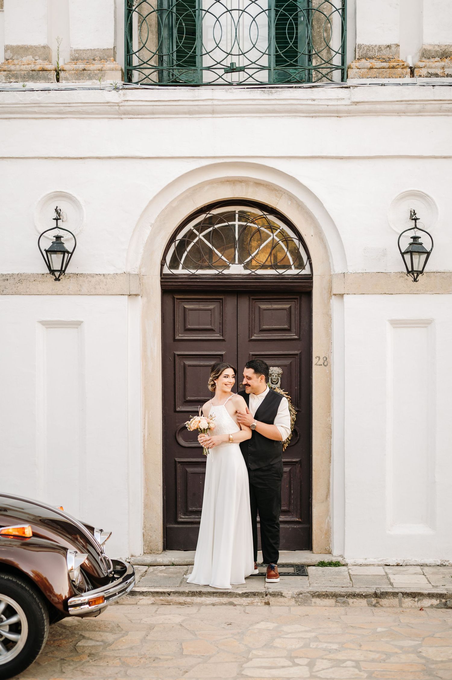 Elegant couple poses in front of white building with ornate dark wooden doors.