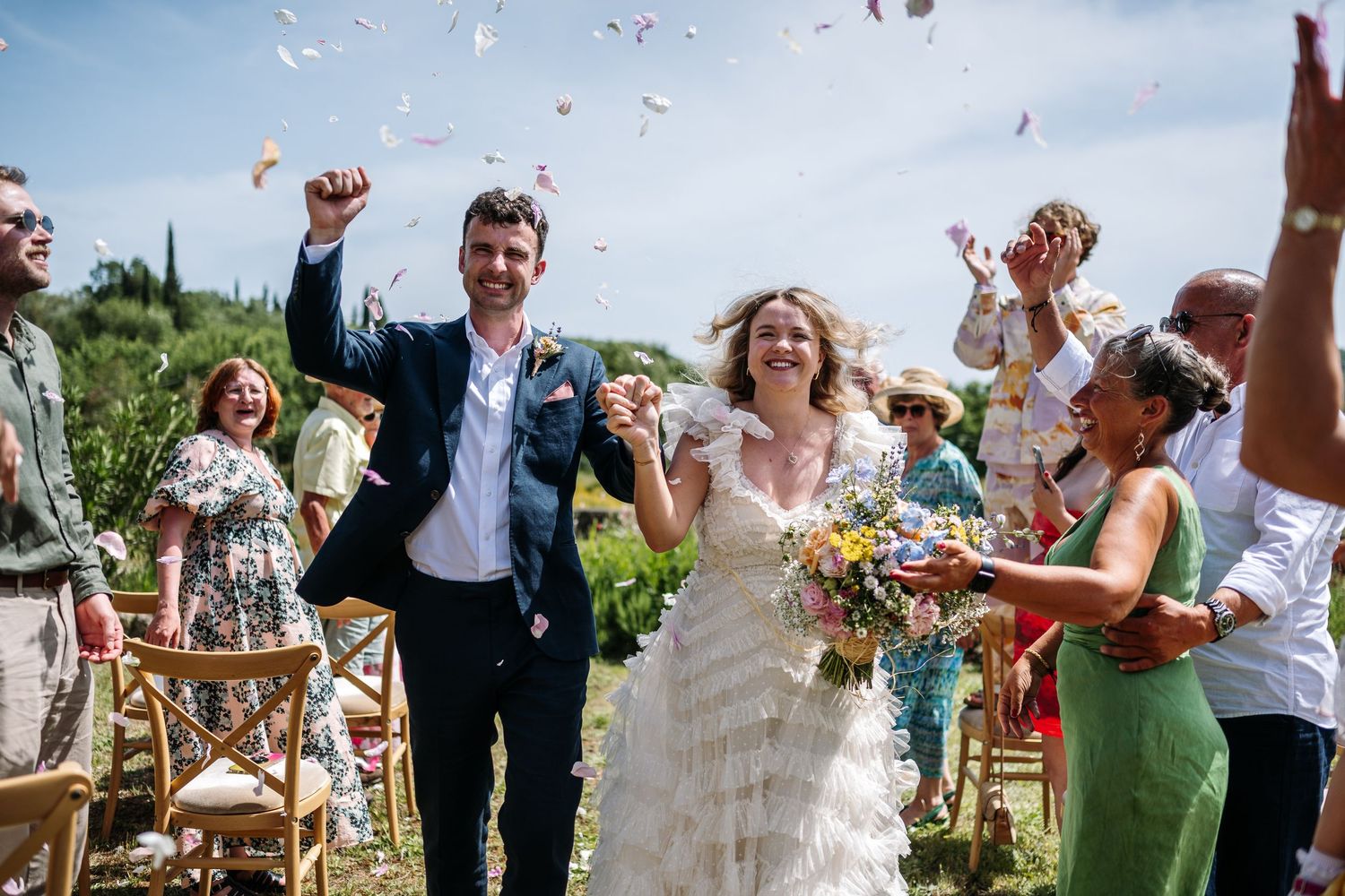 Wedding celebration with guests throwing confetti in a sun-drenched outdoor setting.