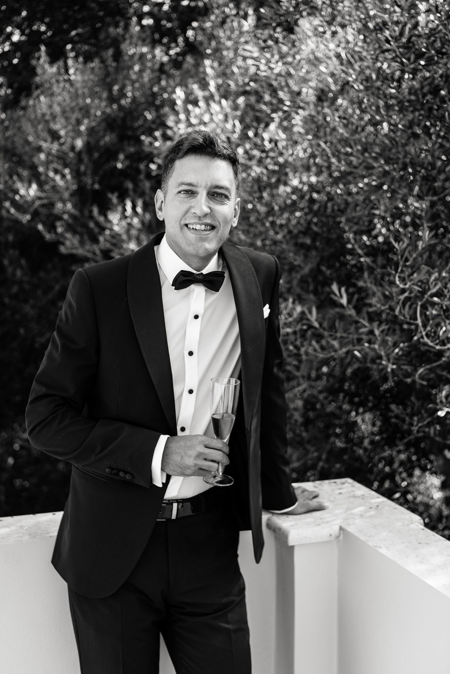 Person in black tuxedo and bowtie leaning on white surface outdoors.