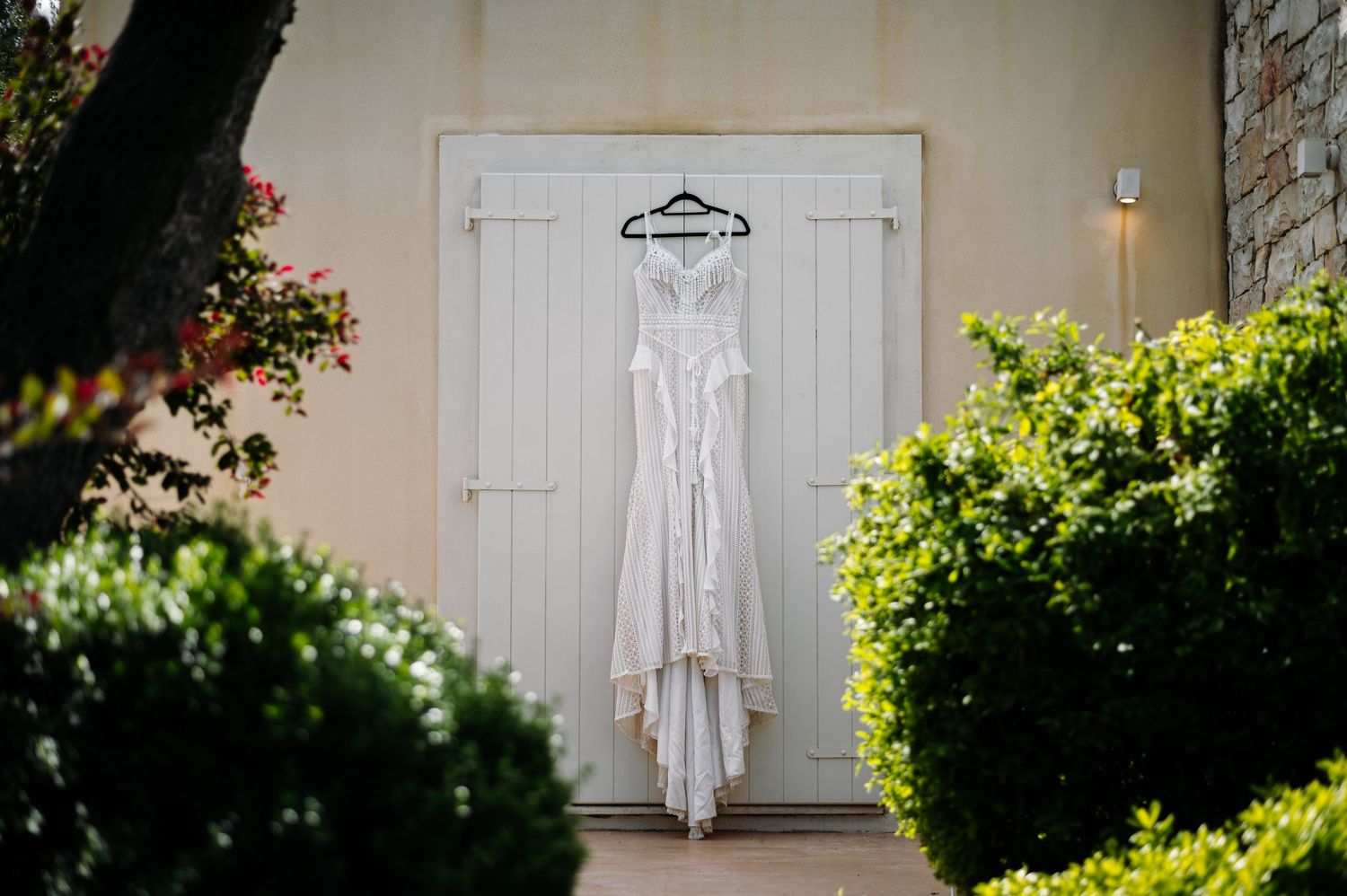 White wedding dress hanging on rustic door surrounded by green bushes.