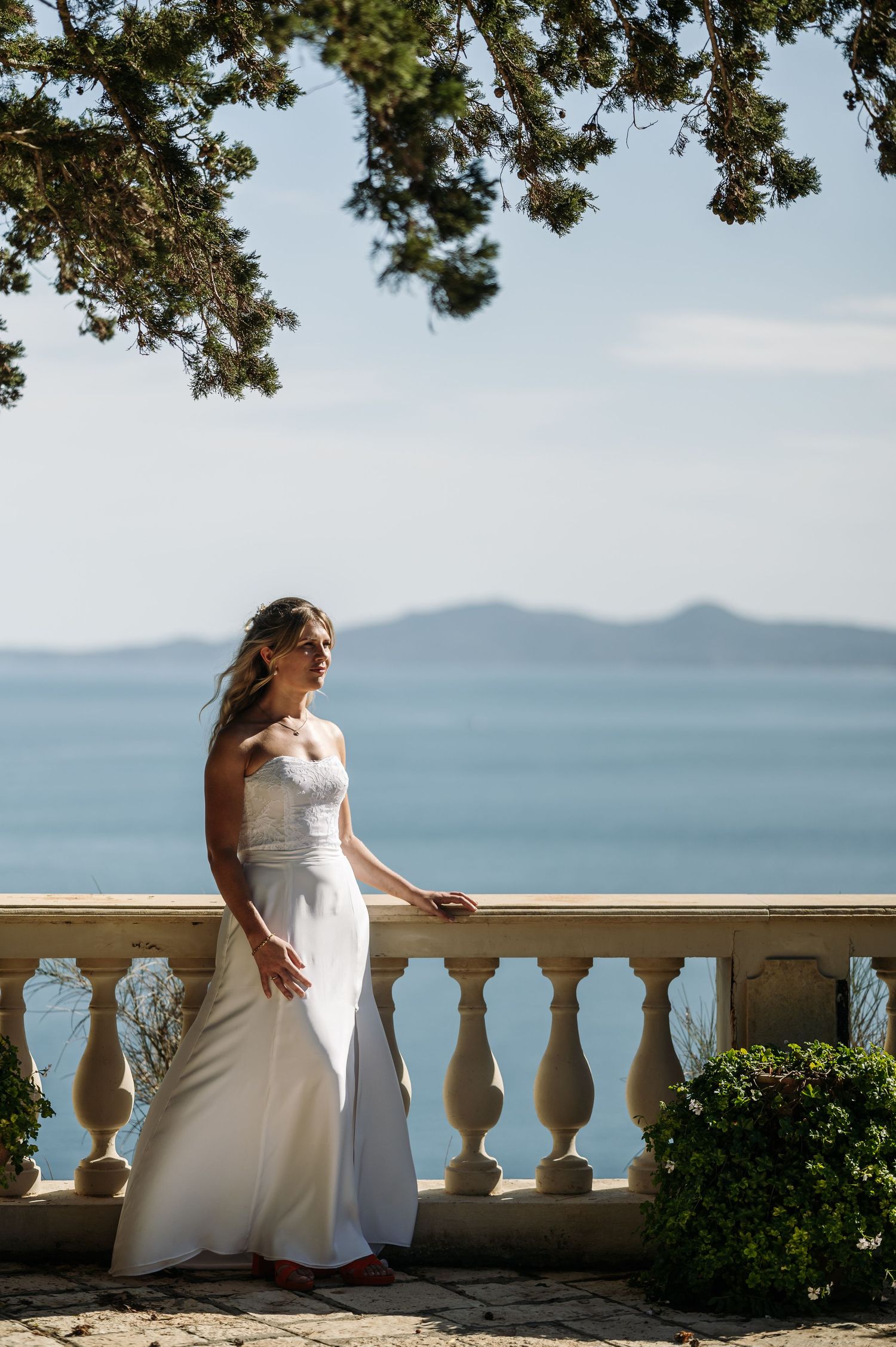 A bride rests on a stone balcony overlooking a serene ocean and mountain landscape.