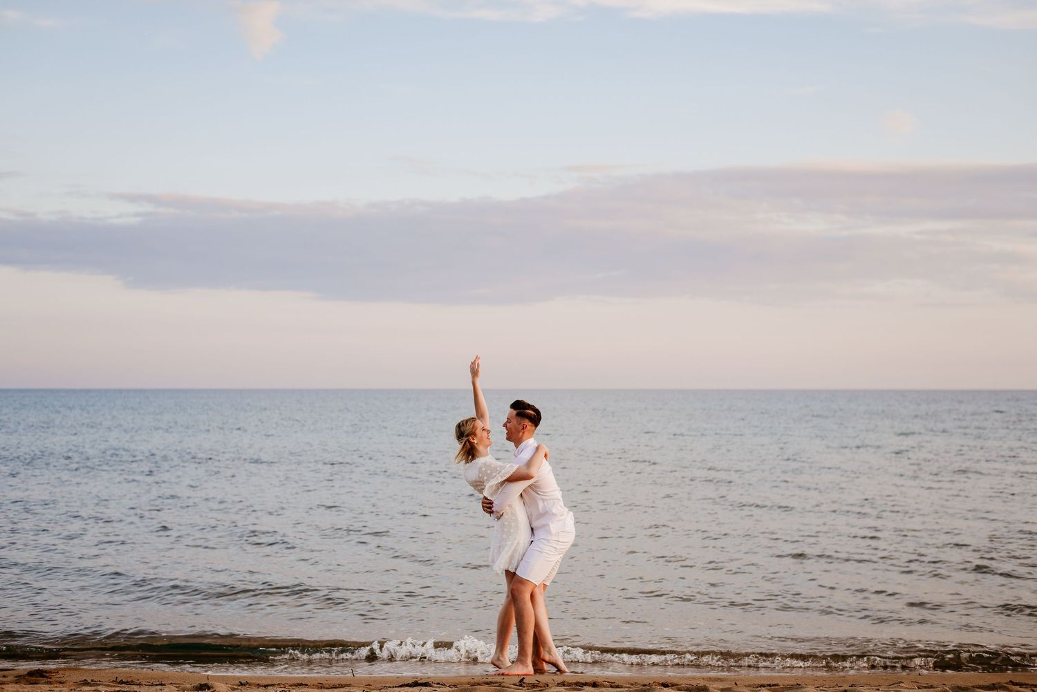 Couple in white clothing dancing joyfully on a beach at sunset.