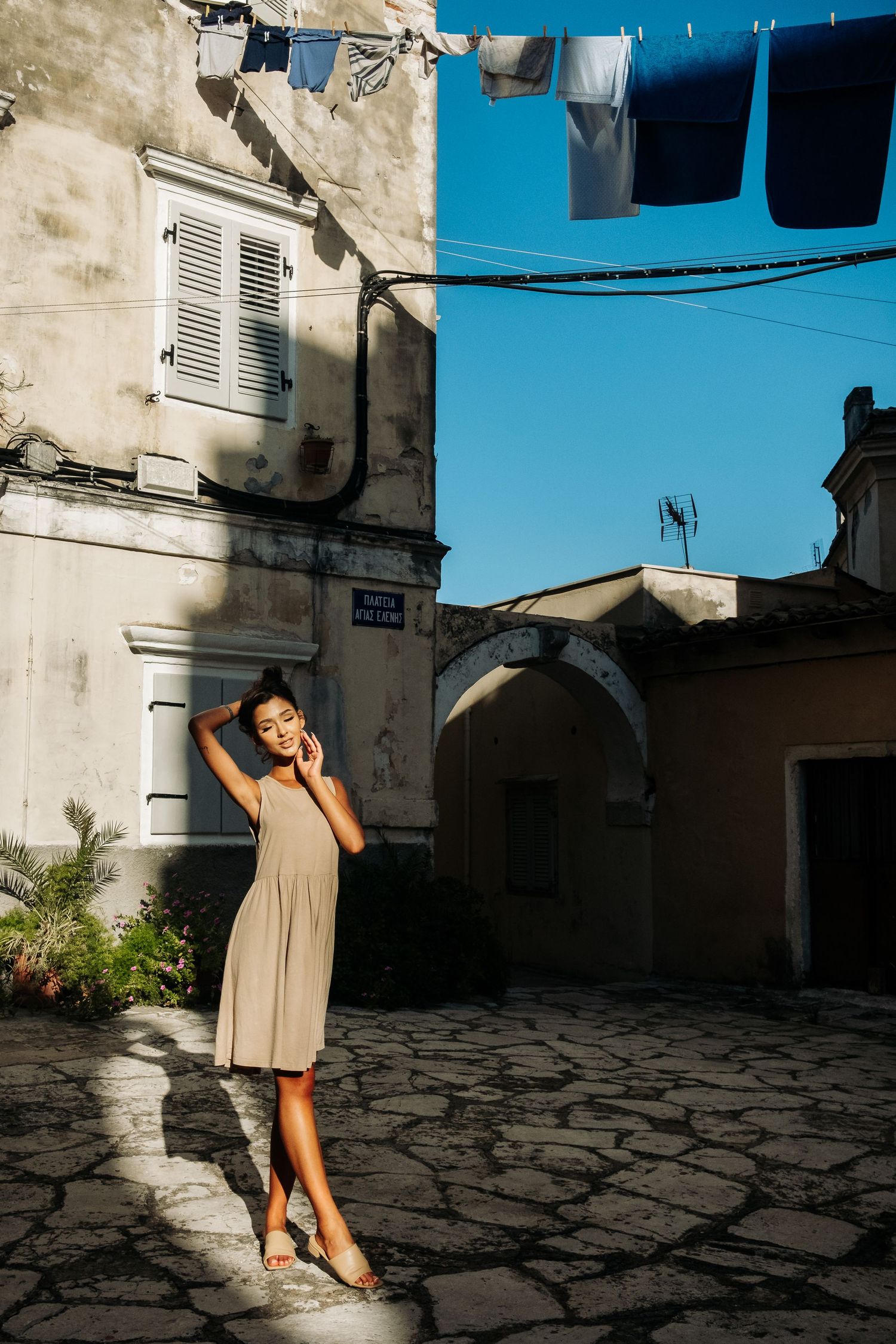 Woman in beige dress dancing in sunlit courtyard with white walls and blue sky.