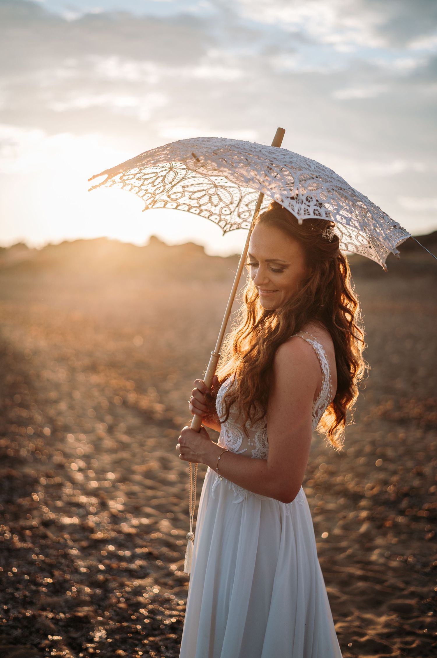 Bride with lace parasol poses on beach during golden hour photography session.