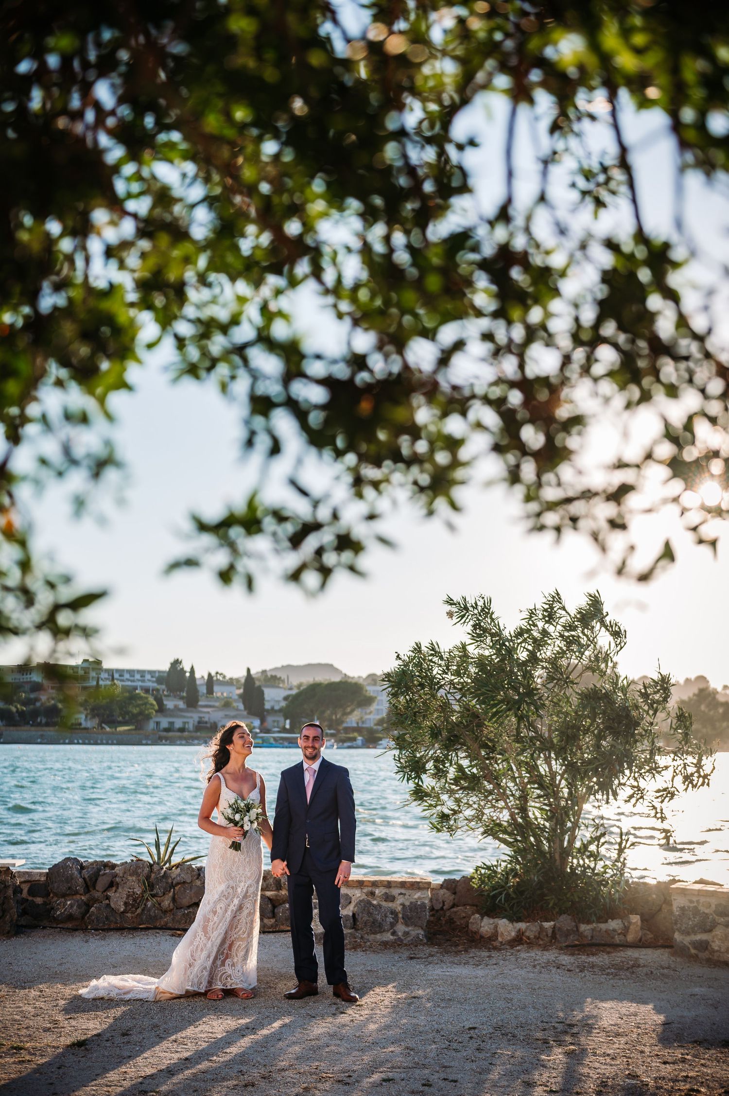 Wedding couple poses along a scenic waterfront with palm trees at sunset.
