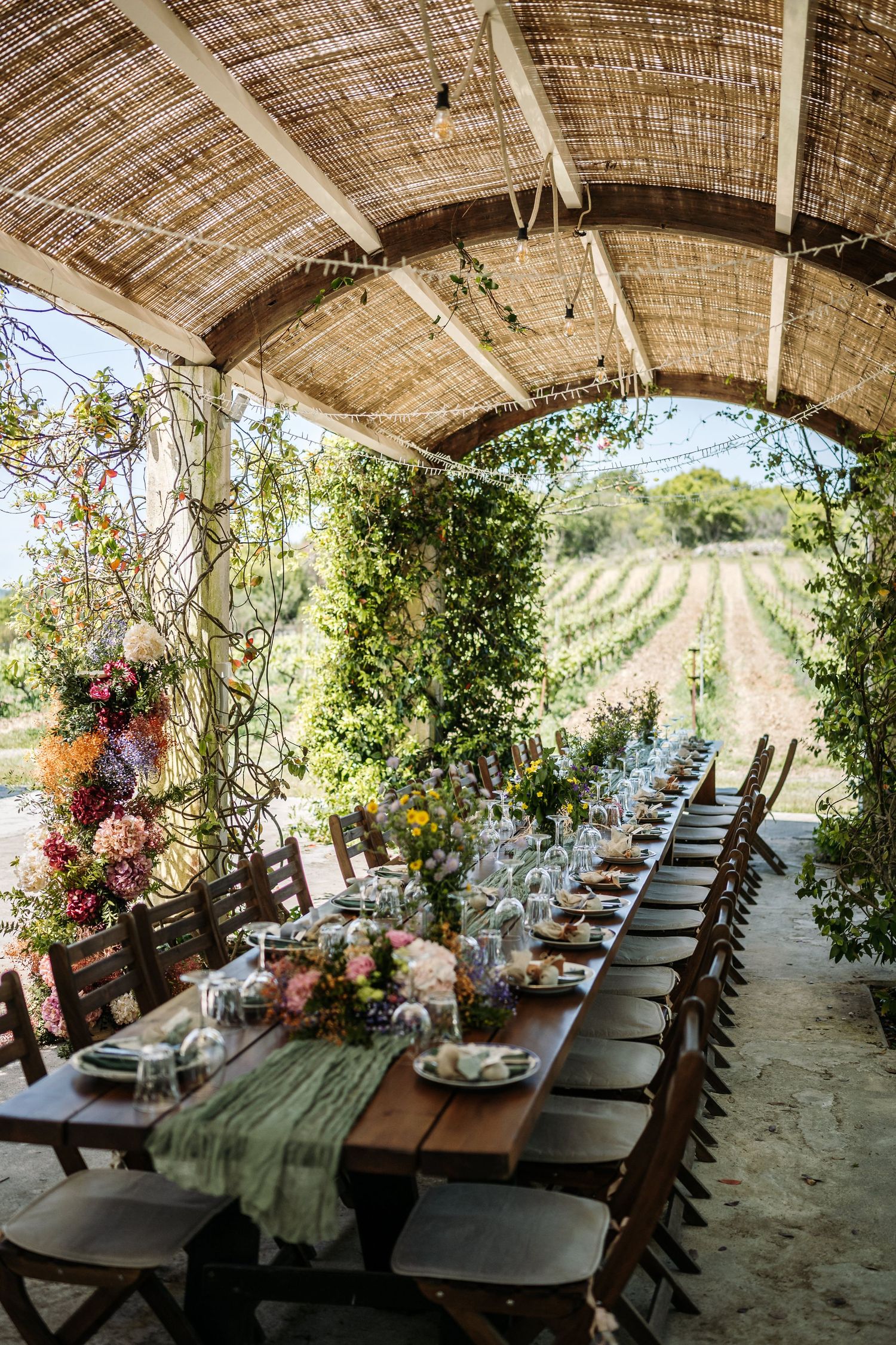 Long rustic dining table set under a covered patio with vineyard views and floral arrangements.