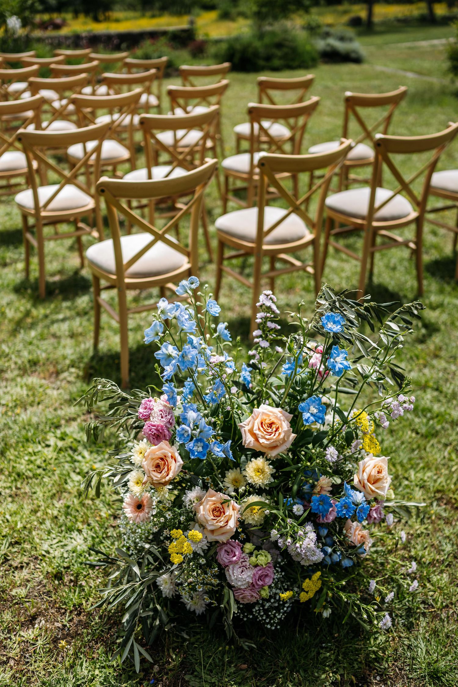 Wooden ceremony chairs arranged on grass with colorful floral arrangements featuring blue and pink blooms.