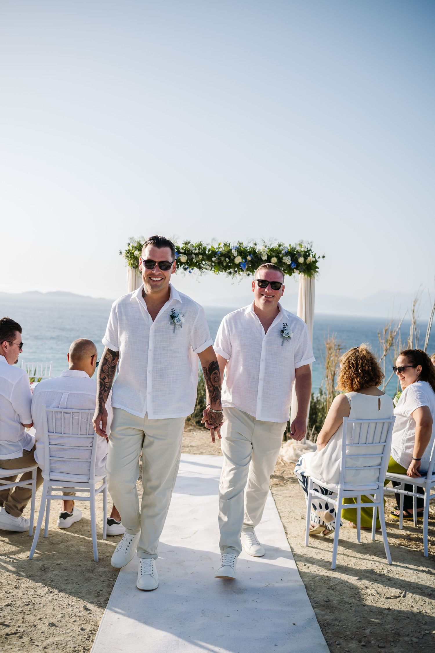 Wedding guests in all-white attire gather at beach ceremony with ocean views.