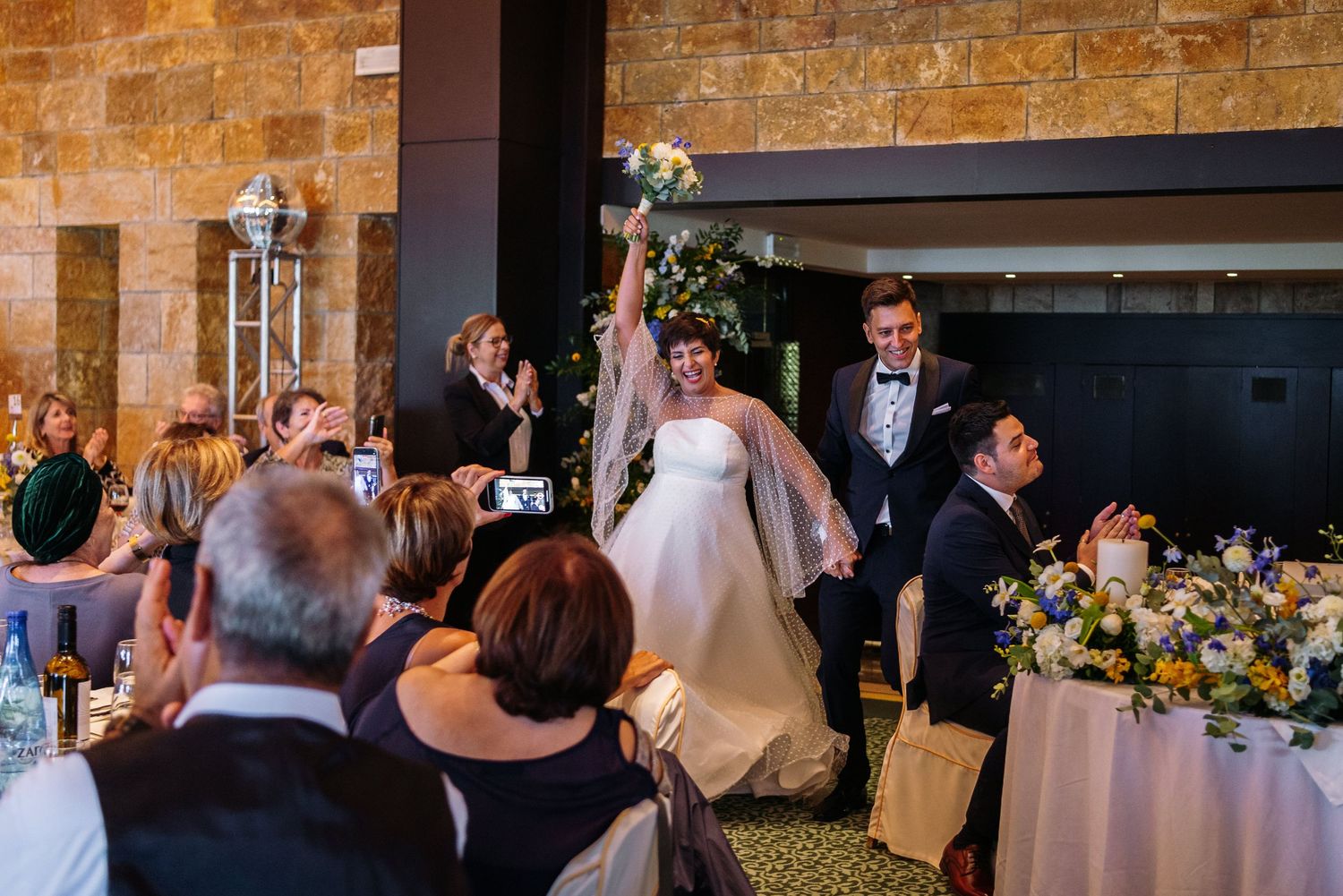 Wedding reception guests celebrate in a rustic brick-walled venue with warm lighting.