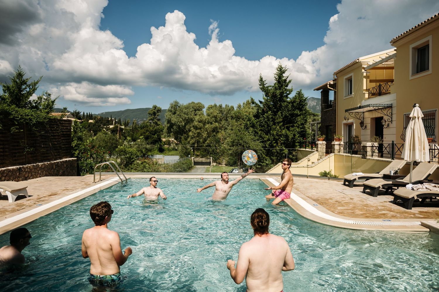 People enjoying a sunny day in a resort swimming pool with blue skies and white clouds.
