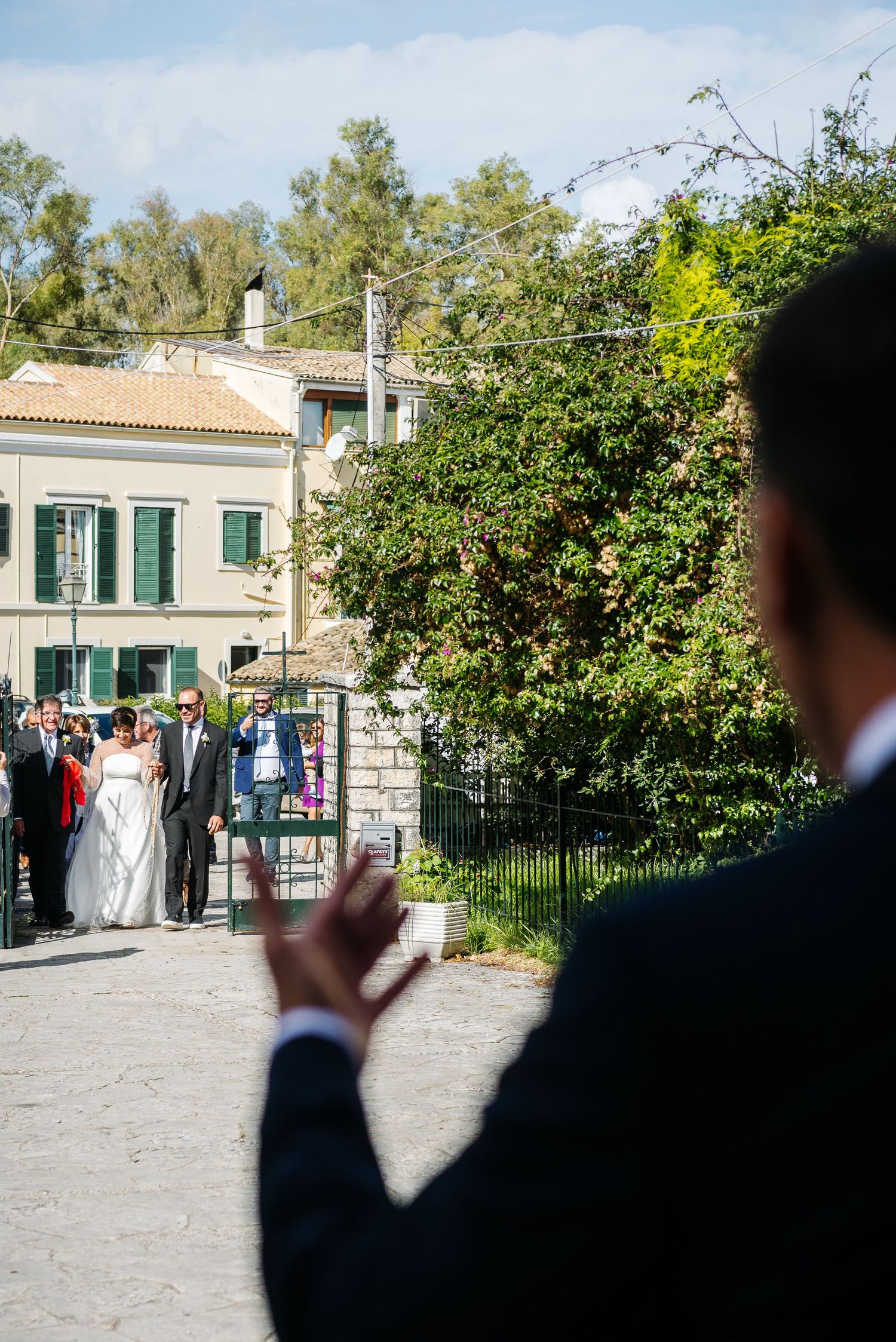 Wedding guests gather outside a Mediterranean-style building with green shutters and lush gardens.