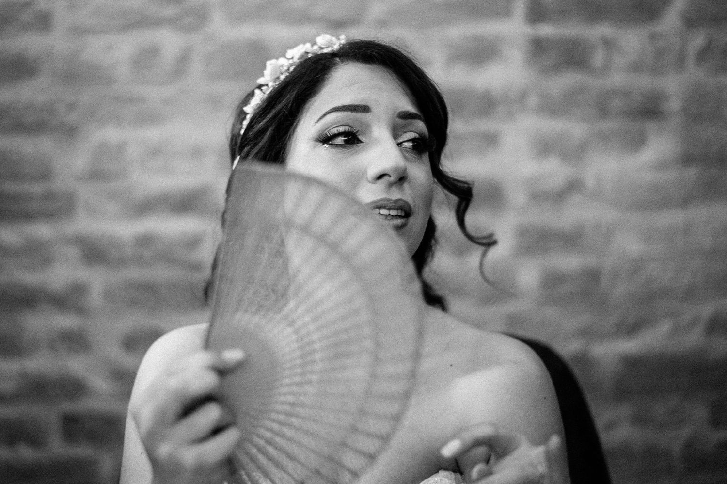 A bride in a white gown poses with a delicate fan against a rustic brick backdrop.