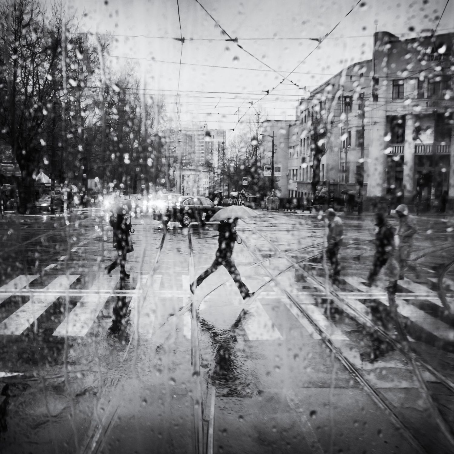 Rain-spattered window view of a city street with blurred figures crossing in the wet weather.