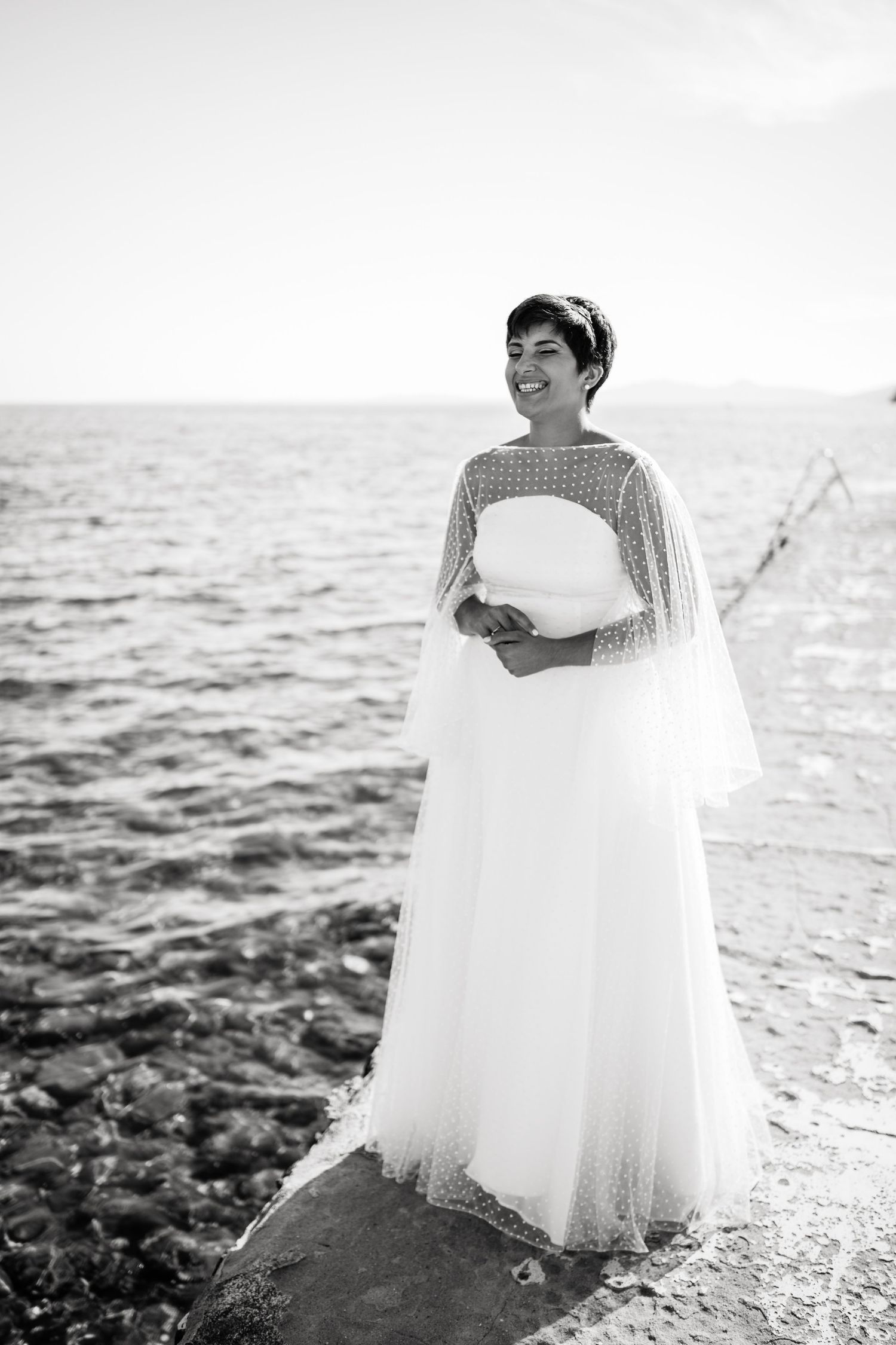 A white wedding dress billows in the breeze along a rocky shoreline at sunset.