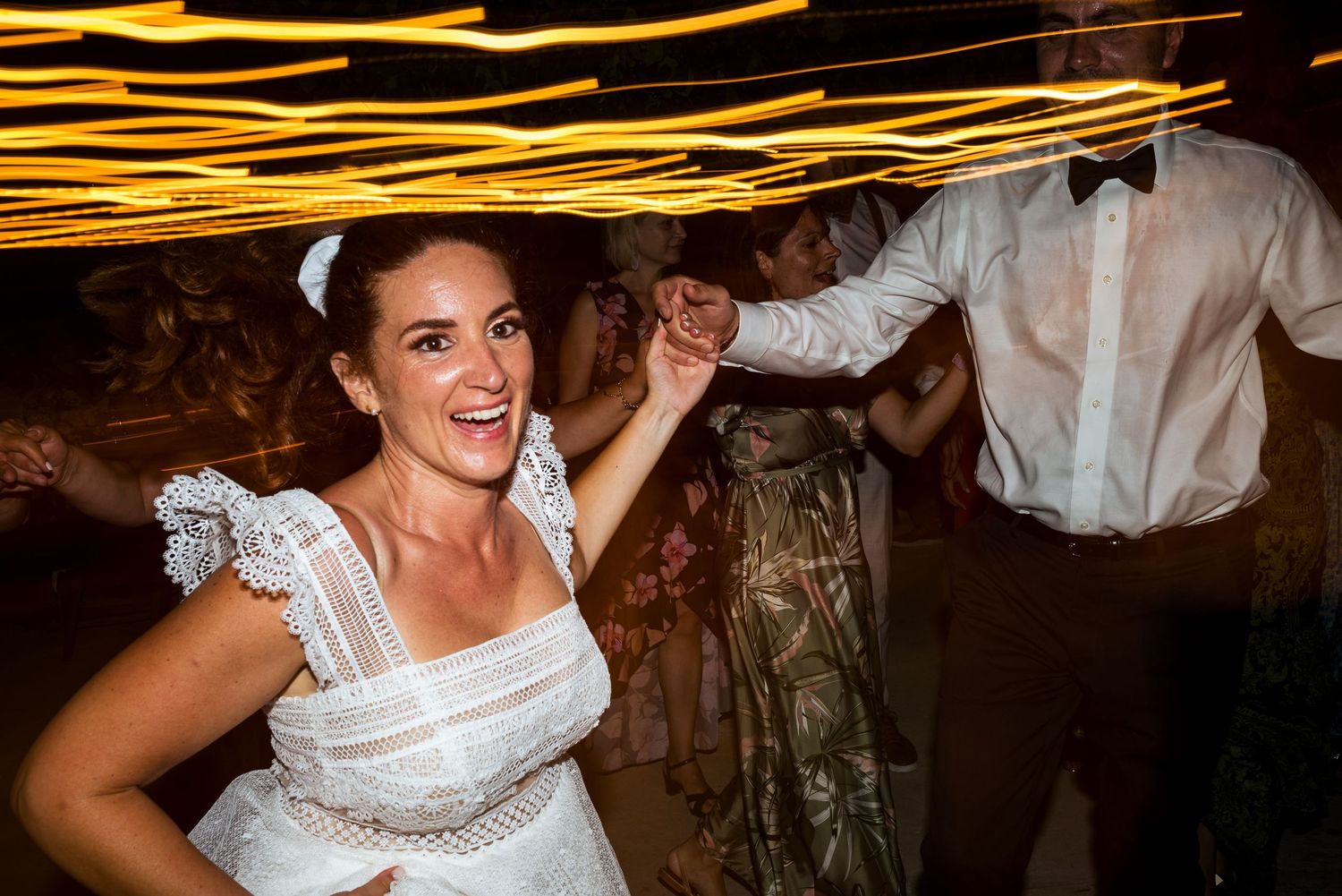 Light trails create glowing patterns above dancers at a nighttime wedding celebration.